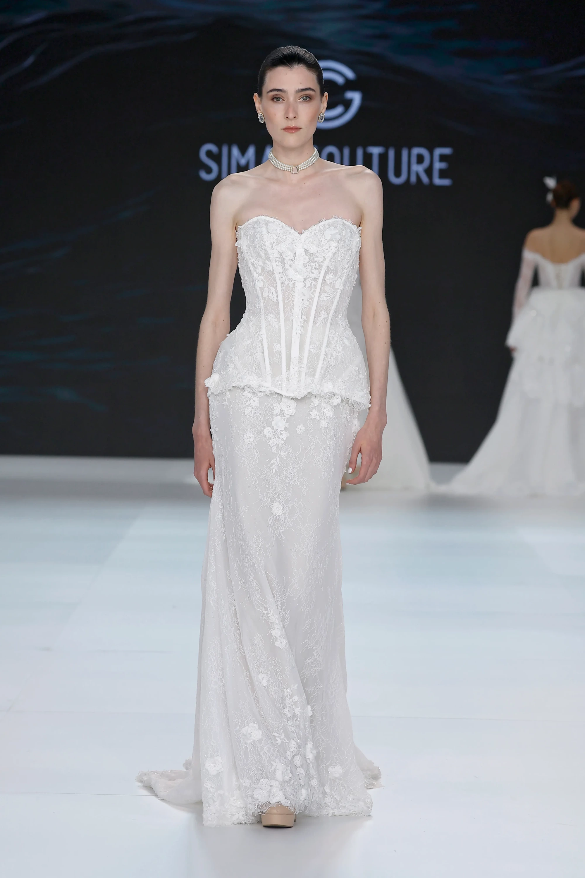 Sima Couture podyumunda, incelikli dantel işlemelerle bezenmiş, korse detaylı ve zarif dekolteli, beyaz, straplez bir gelinlik ile inci gerdanlık takan model, 2027 gelinlik trendleri arasında yer alan modern romantizmi ve Viktoryen dokunuşları yansıtan kabartmalı 3D çiçek aplikeli etek kısmıyla dikkat çekiyor.
