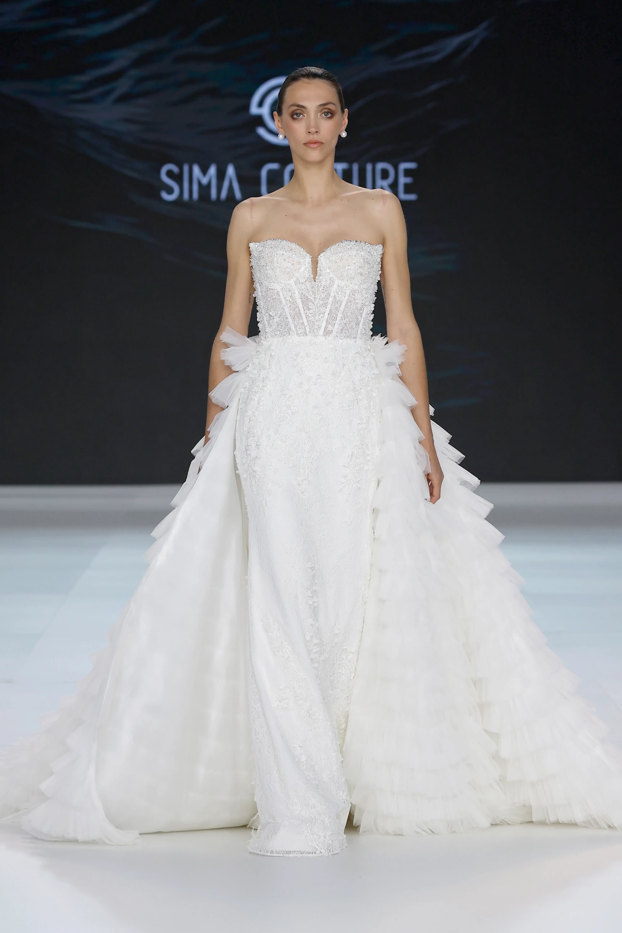 Sima Couture podyumunda, straplez, zarif işlemeli korsajı ve belden itibaren kat kat, fırfırlı tüllerle dramatik bir hacim kazanan beyaz etekli bir gelinlik modeli sergileyen manken, 2027 gelinlik trendleri içinde modern romantizmi yansıtan bir görünüm sunuyor.