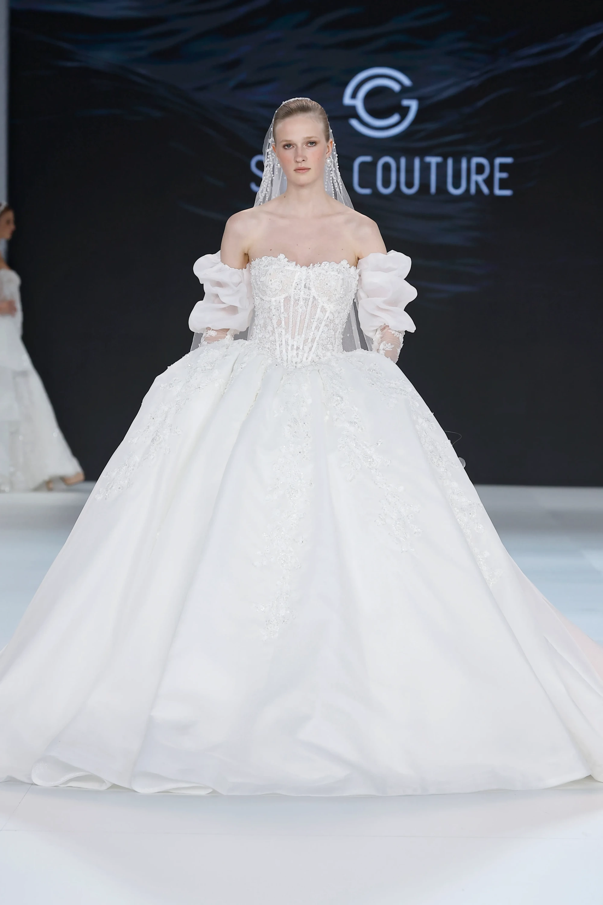 Sima Couture defilesinde podyumda duran bir model, 2027 gelinlik trendlerini yansıtan, omuzları açıkta bırakan korse kesimli üstü, hacimli puf kolları ve zarif dantel detaylarla bezenmiş, prenses siluetindeki bembeyaz ihtişamlı bir gelinlik içinde; yüzünü çerçeveleyen incili duvağı ile göz kamaştırıyor.