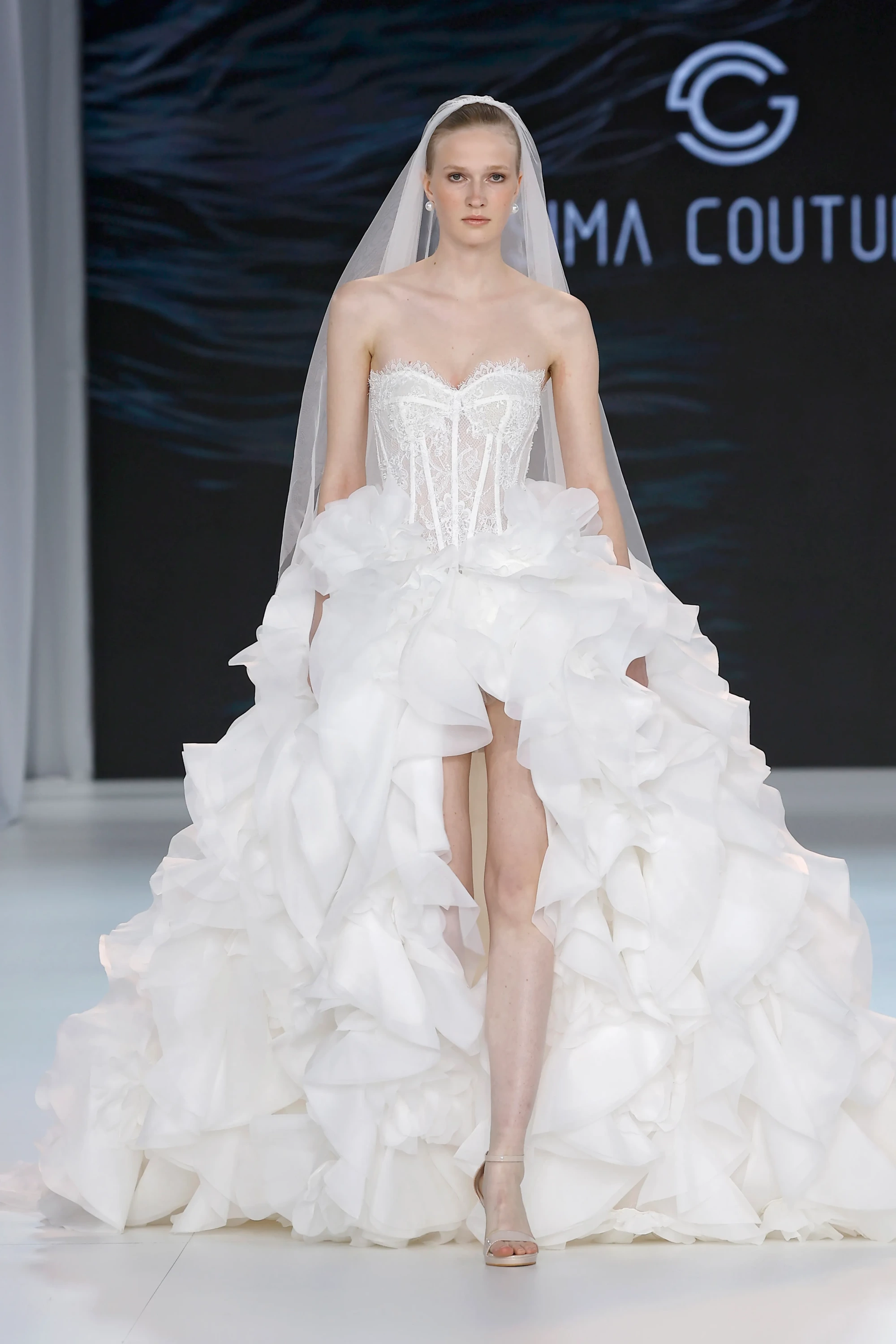 Sima Couture imzalı, straplez dantel korse büstiyerli ve balerin etkisini yansıtan kat kat, hacimli fırfırlı eteği yüksek yırtmaçla modern bir dokunuş kazanan beyaz gelinliği, sade tül duvak ve inci küpelerle sergileyen bir model, 2027 gelinlik trendlerini podyumda gözler önüne seriyor.