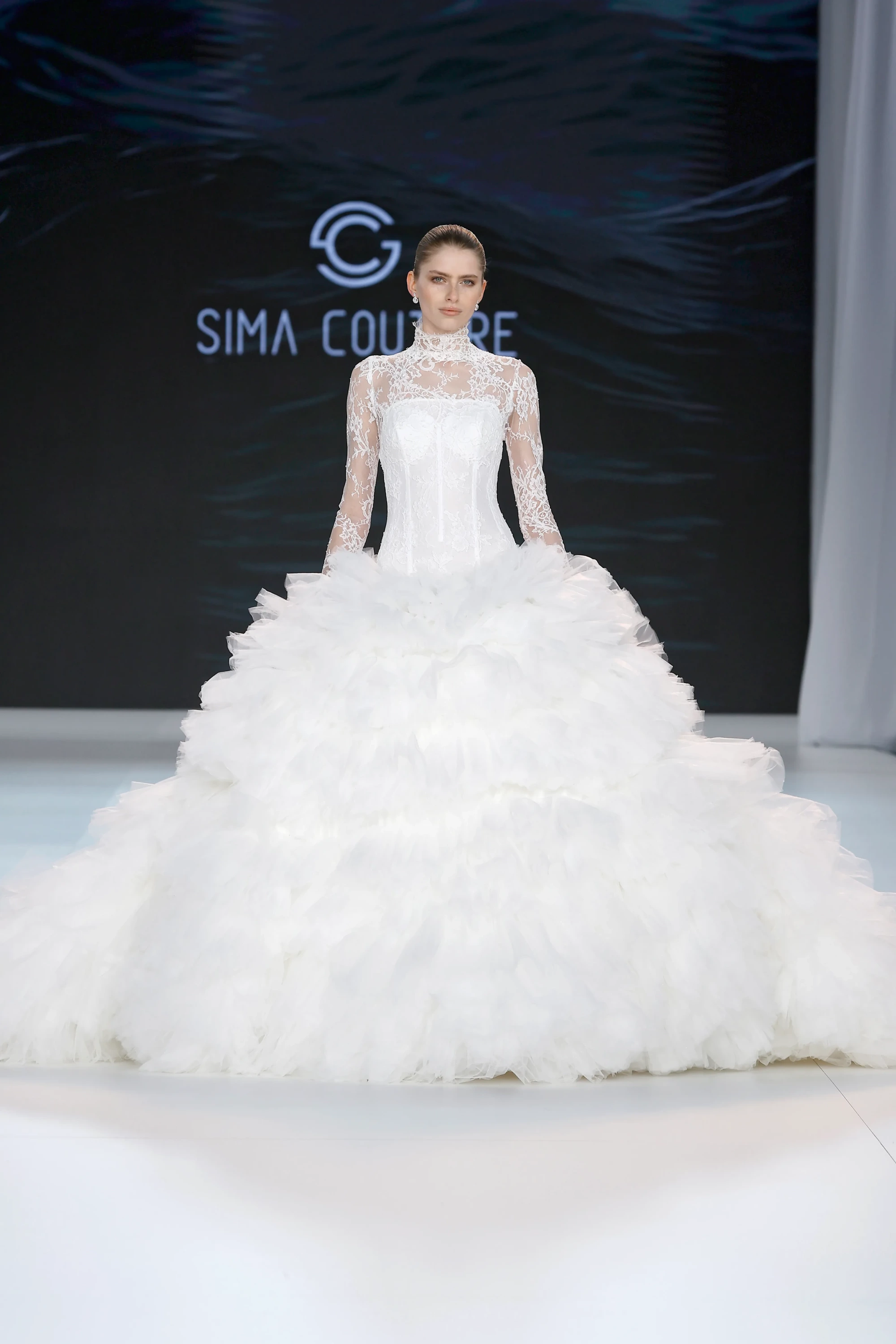 Podyumda duran bir model, yüksek dantel yakalı ve uzun transparan dantel kollu, katmanlı ve fırfırlı tüllerden oluşan inanılmaz hacimli eteğiyle göz alıcı bir beyaz Sima Couture gelinlik sunuyor, bu tasarım 2027 gelinlik trendlerinin zarif balerin etkisini gözler önüne seriyor.