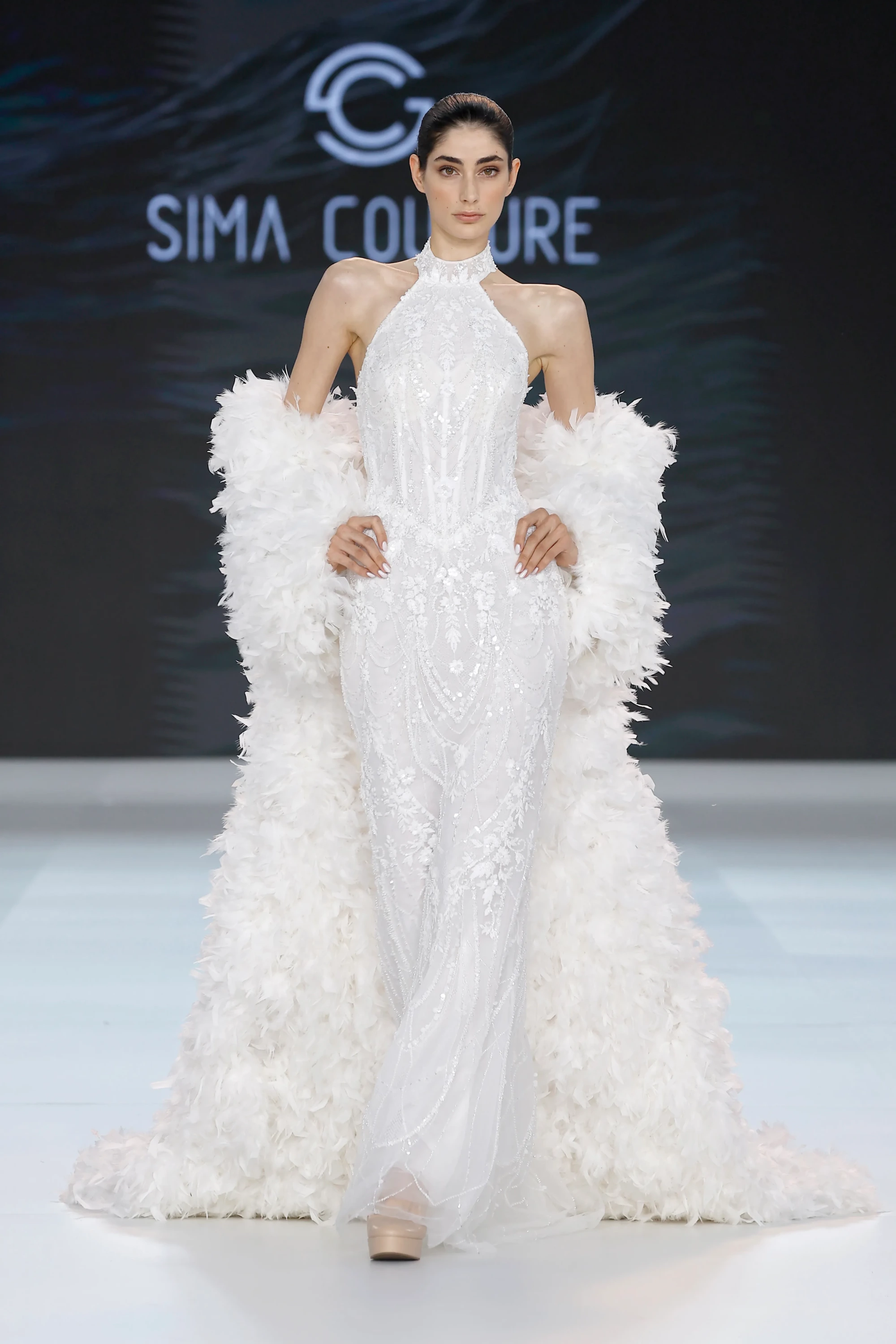 Sima Couture defilesinde, yüksek yakalı, tamamı göz alıcı parıltılı işlemelerle süslü balık formundaki beyaz bir gelinlik giyen model, omuzlarından yere kadar uzanan gösterişli beyaz tüy pelerinle 2027 gelinlik trendlerini sergiliyor.