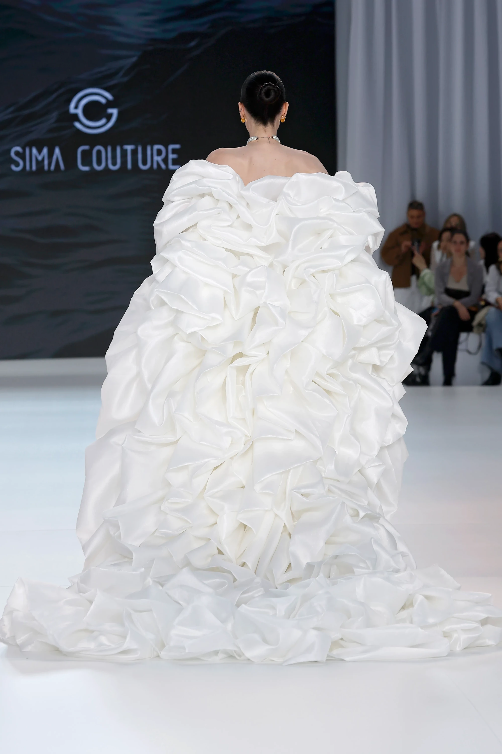 Sima Couture defilesinde, sırtı dönük bir model, markanın 2027 gelinlik trendlerine uygun, yoğun fırfırlı ve drapeli, hacimli ekru rengi bir gelinlik içinde yürüyor; podyumun arka planında Sima Couture logosu dikkat çekiyor.