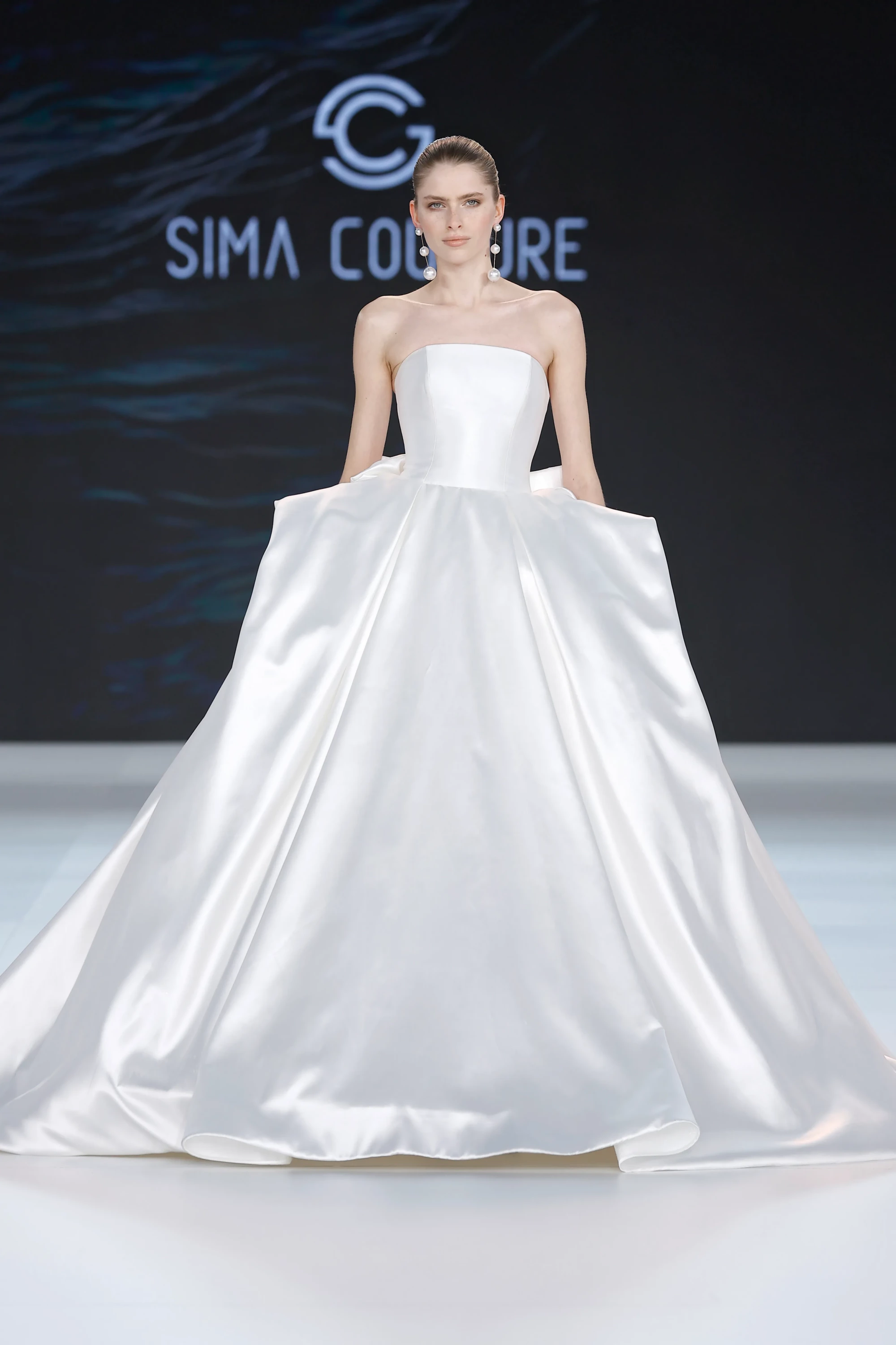 Podyumda Sima Couture imzalı, straplez kesimli, parlak saten kumaştan tasarlanmış hacimli eteğiyle dikkat çeken bu zarif gelinlik, 2027 gelinlik trendlerinin zamansız ve romantik yorumlarından birini sergiliyor.