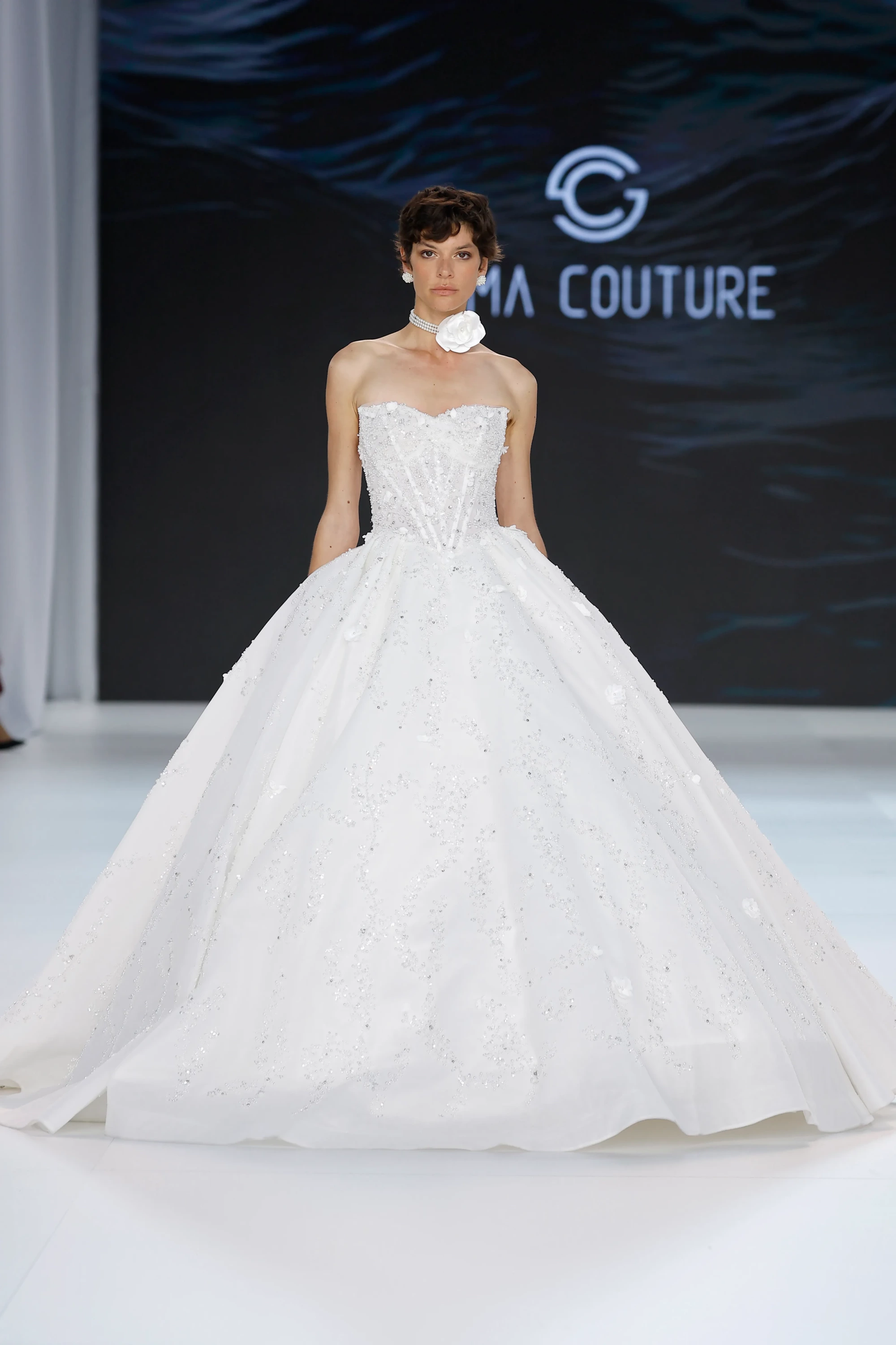 Sima Couture defilesinden beyaz, straplez, korsajlı ve kabarık etekli, baştan aşağı pırıltılı işlemelerle bezeli bu prenses kesim gelinlik, incili yakasındaki büyük beyaz çiçek detayıyla 2027 gelinlik trendlerini yansıtan iddialı bir silüet sunuyor.