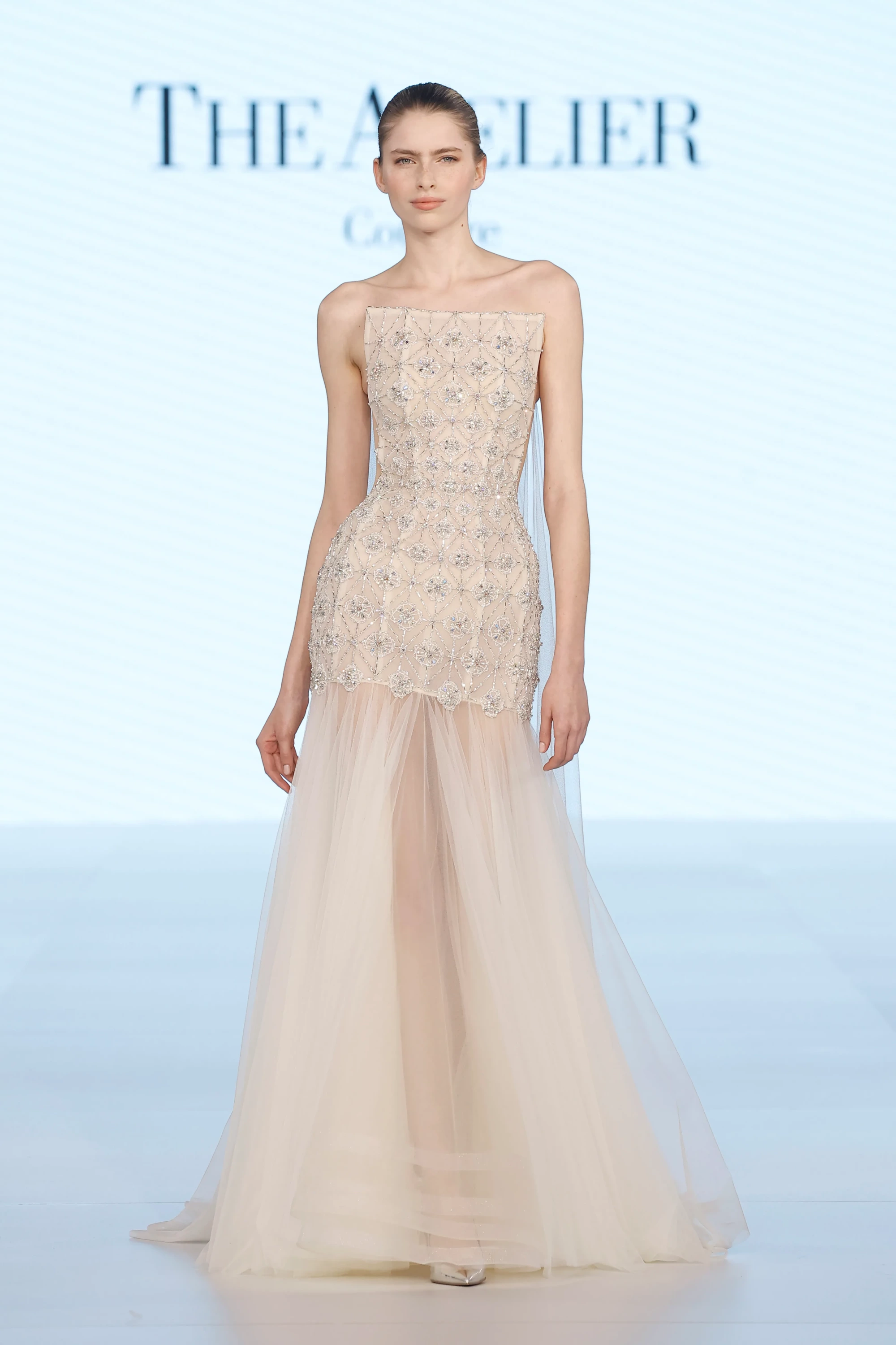 Podyumda duran model, The Atelier by Prof. Jimmy Choo'nun ustalıklı terziliğini yansıtan, üst kısmı gümüş kristaller ve boncuklarla işlenmiş, geometrik desenlere sahip straplez, açık bej bir gelinlik sergiliyor; katmanlı şeffaf tül eteği ve ışıltılı detaylarıyla bu tasarım, 2027 gelinlik trendleri arasında öne çıkan zarafeti ve akışkanlığı mükemmelen temsil ediyor.
