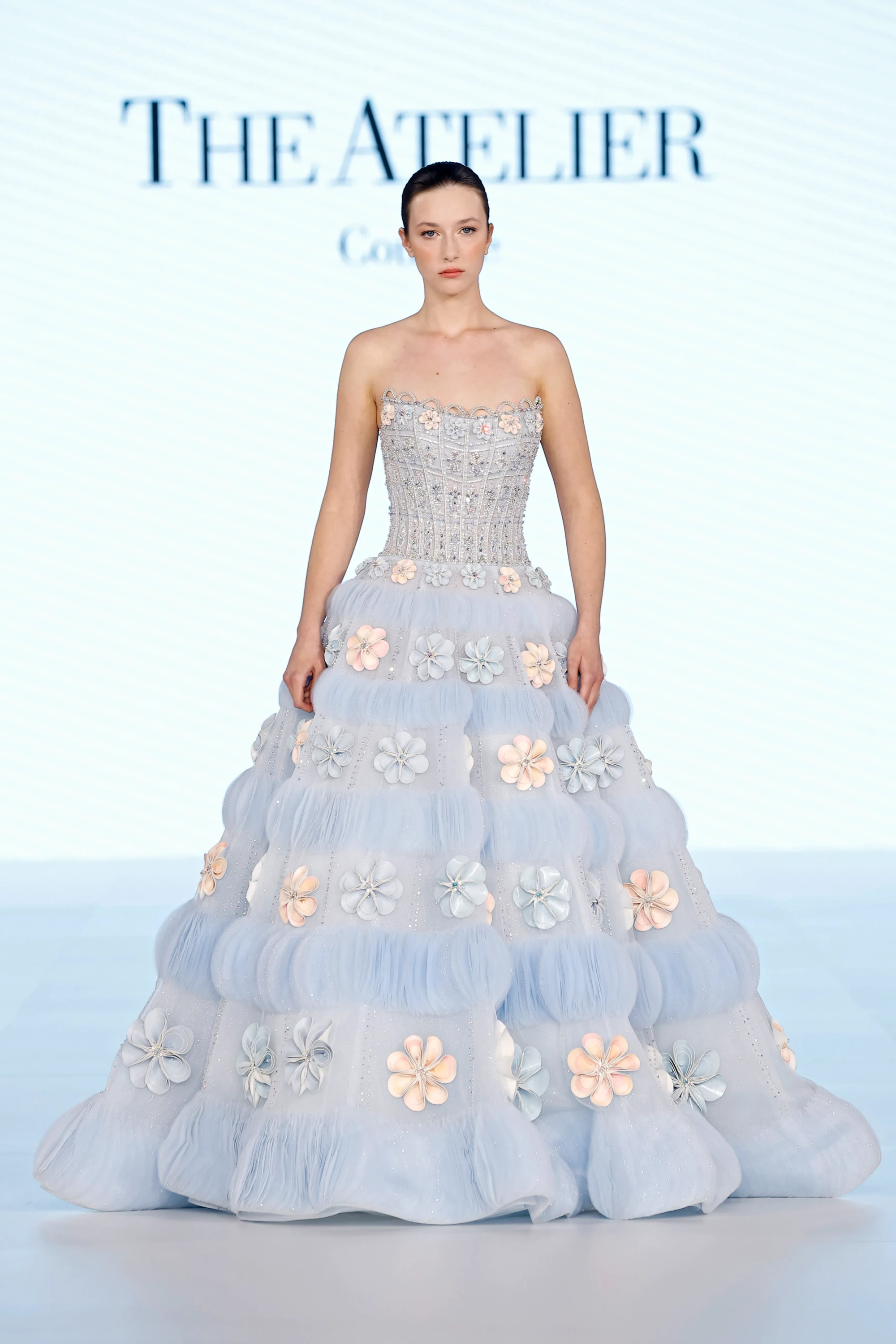 Model, podyumda The Atelier by Prof. Jimmy Choo koleksiyonundan, işlemeli korsajı ve katmanlı hacimli tül eteği üzerinde açık mavi ve şeftali tonlarında 3D çiçek aplikeleri ile süslenmiş, 2027 gelinlik trendlerini yansıtan, parıldayan bir gelinlik sergiliyor.