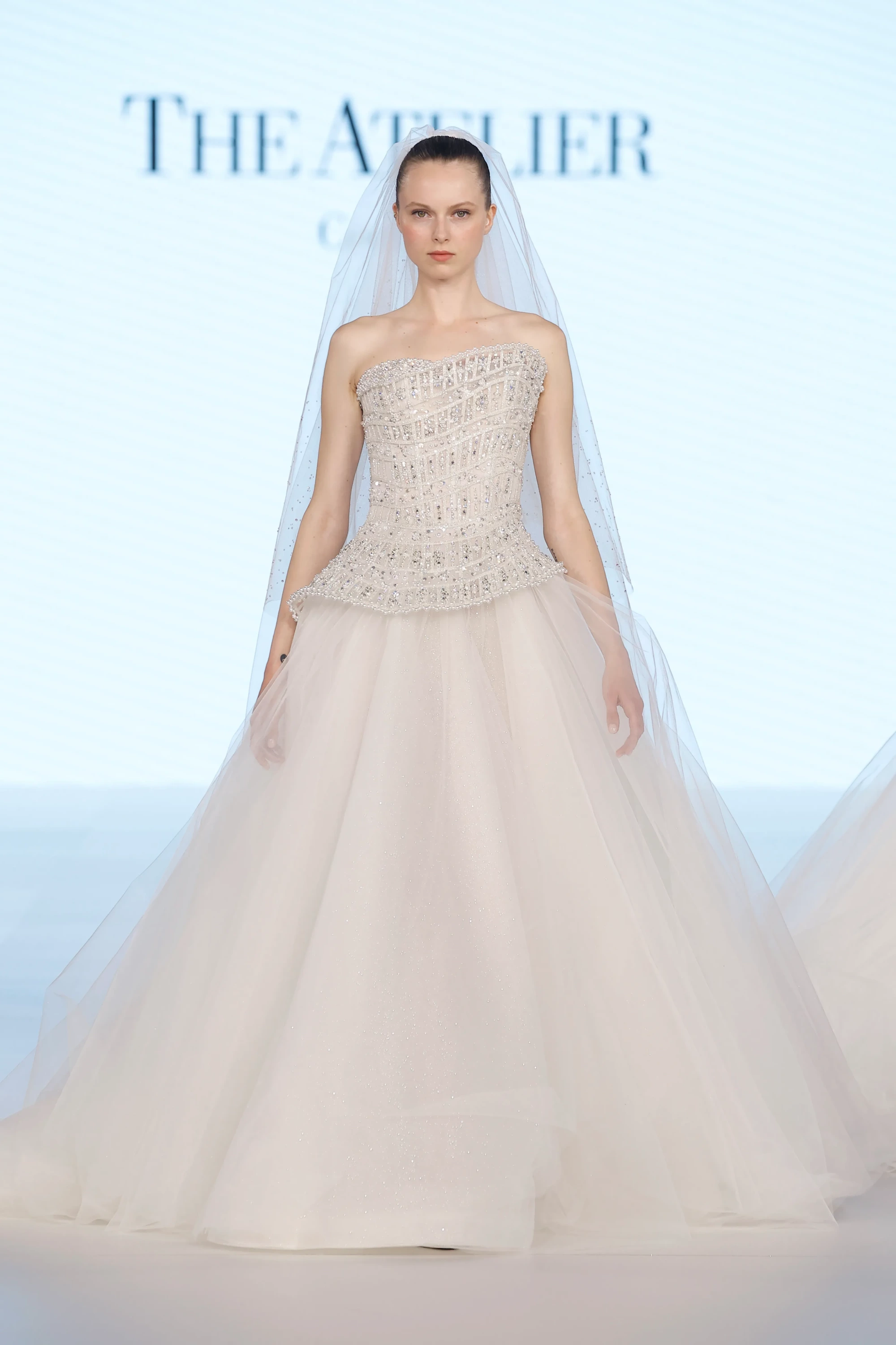 Podyumda duran model, The Atelier by Prof. Jimmy Choo imzalı, üst kısmı gümüş payet ve kristal işlemeli, güçlü korsajlı ve yıldız ışıltısını anımsatan katmanlı, hacimli tül eteğe sahip zarif bir 2027 gelinlik modeli sergiliyor.