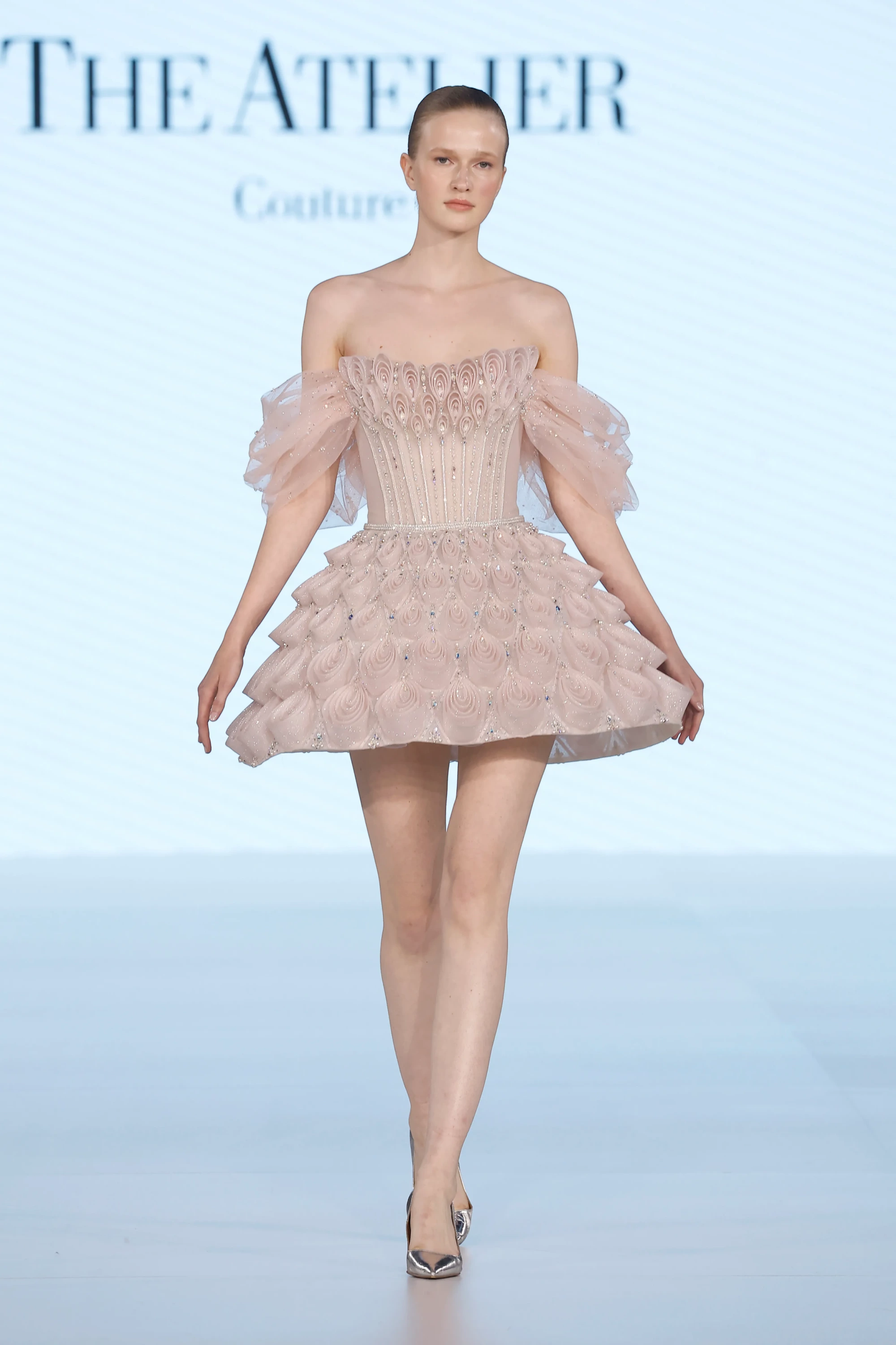 The Atelier by Prof. Jimmy Choo'nun 2027 İlkbahar koleksiyonundan, podyumda yürüyen model; ışığı yansıtan pudra pembe tonlarındaki, omuzları açık, pul benzeri katmanlı işlemeler ve Swarovski kristallerle süslü korsajıyla dikkat çeken, kısa ve hacimli eteğiyle hafifliği vurgulayan, tül fırfırlı kollara sahip, romantik ve modern gelinlik trendlerine uygun zarif bir tasarım sergiliyor.