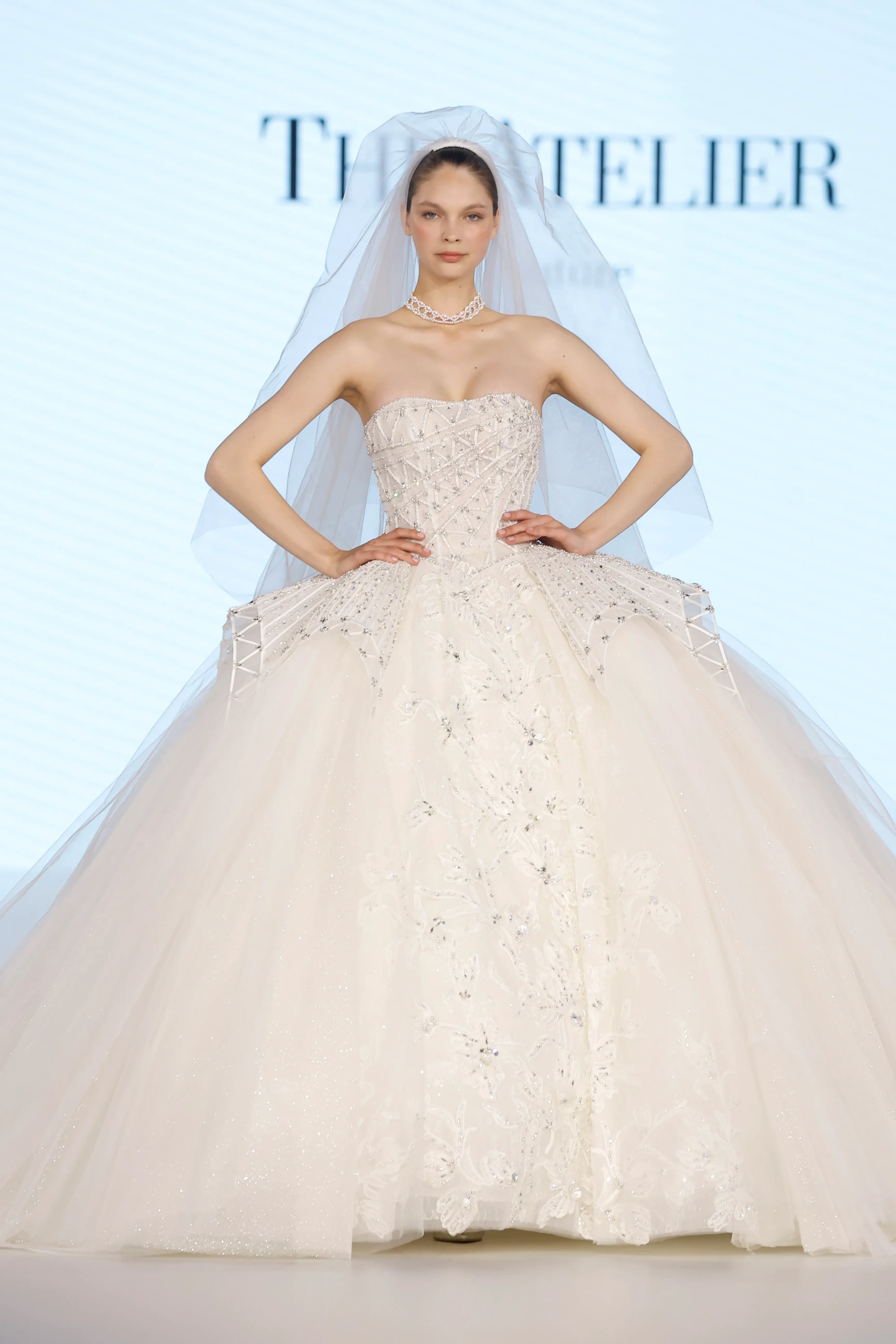 The Atelier markasının 2027 Gelinlik Trendleri koleksiyonundan, güçlü korsajlı, geometrik kristal işlemelerle bezeli üst kısmı ve alt kısmı ince nakışlı, katmanlı ışıltılı tüllerden oluşan hacimli eteğiyle dikkat çeken, inci kolyeli ve zarif duvaklı bir gelinlik podyumda sergileniyor.
