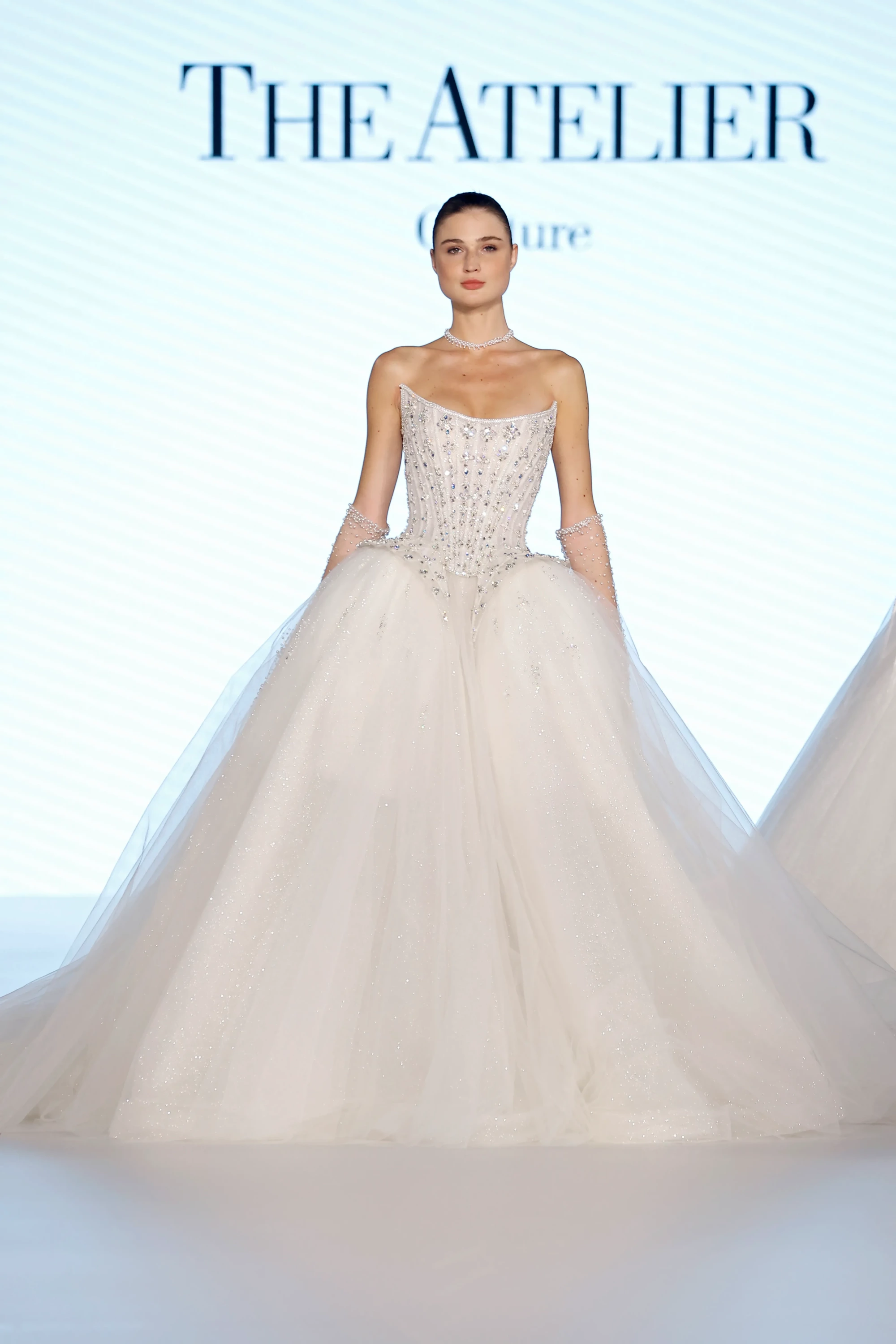 Bir podyumda, The Atelier yazısı önünde duran model, işlemeli korsajlı ve parıltılı katmanlı tüllerden oluşan kabarık etekli, uzun, straplez, kırık beyaz bir gelinlik giyiyor; inci detaylı gerdanlık ve transparan eldivenleriyle bu 2027 gelinlik trendi öne çıkıyor.