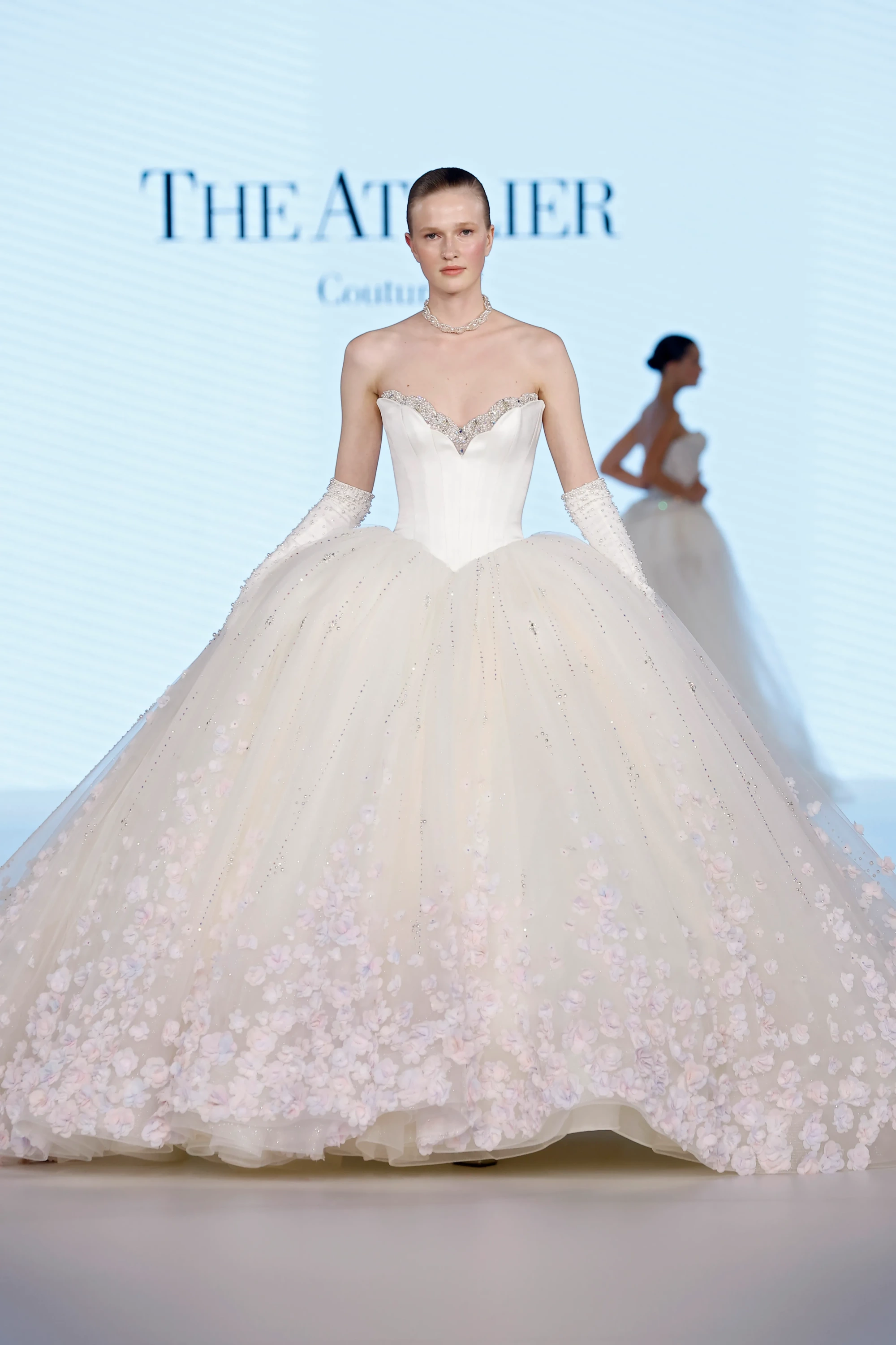 The Atelier markasının bir defilesinde, kristallerle süslü kalp yaka korsajı ve alt eteklerine doğru pembe üç boyutlu çiçek aplikeleriyle bezeli katmanlı, hacimli tül eteği olan zarif bir gelinlik giyen model, uzun inci işlemeli eldivenleri ve kolyesiyle 2027 gelinlik trendlerini podyuma taşıyor.