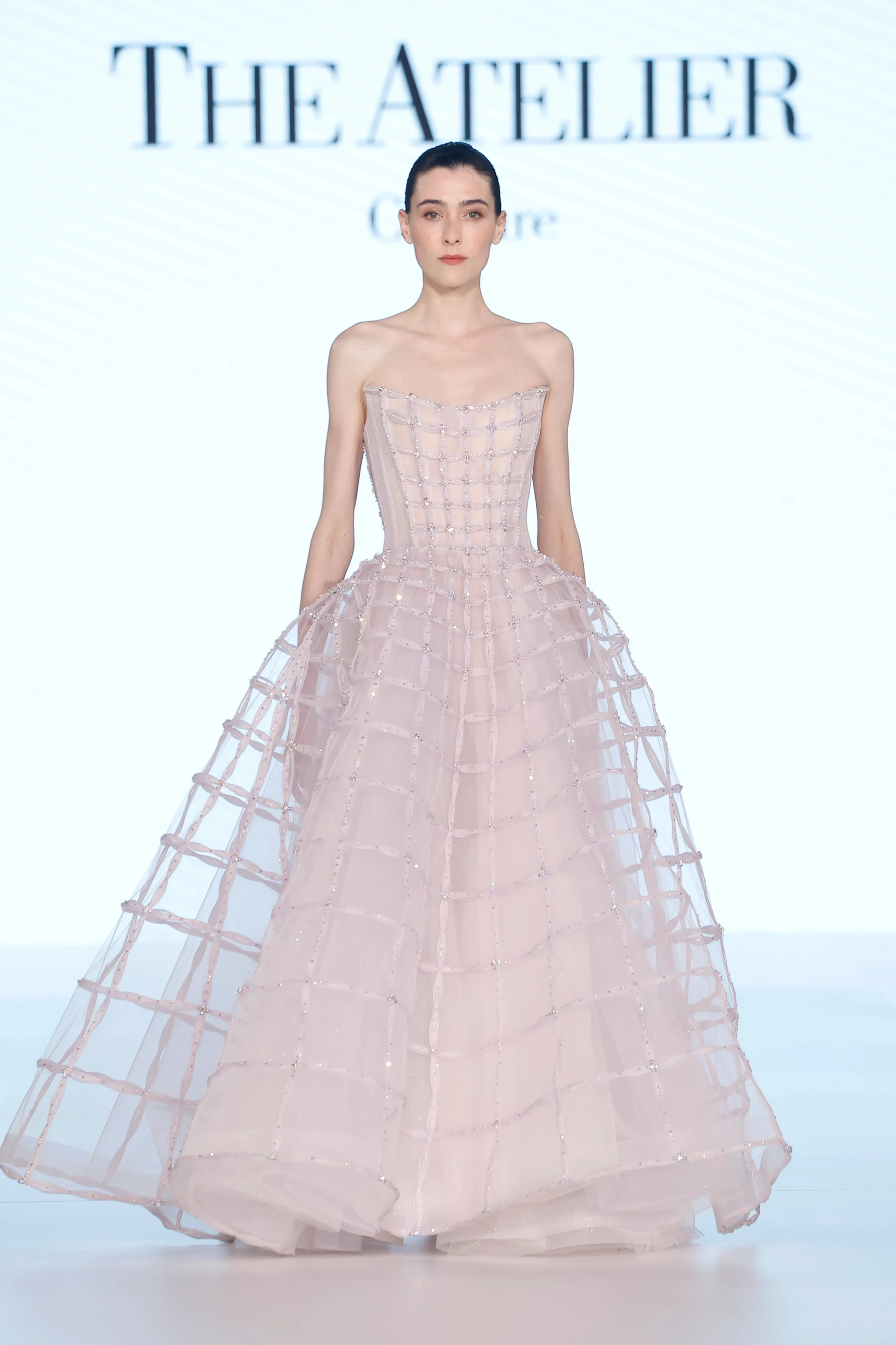 The Atelier by Prof. Jimmy Choo'nun 2027 ilkbahar gelinlik koleksiyonundan podyumda duran model, straplez, güçlü korsajlı, ışıltılı Swarovski kristallerle işlenmiş kafes detaylı, hafif hacimli ve katmanlı, soluk pembe bir gelinlik giyiyor.
