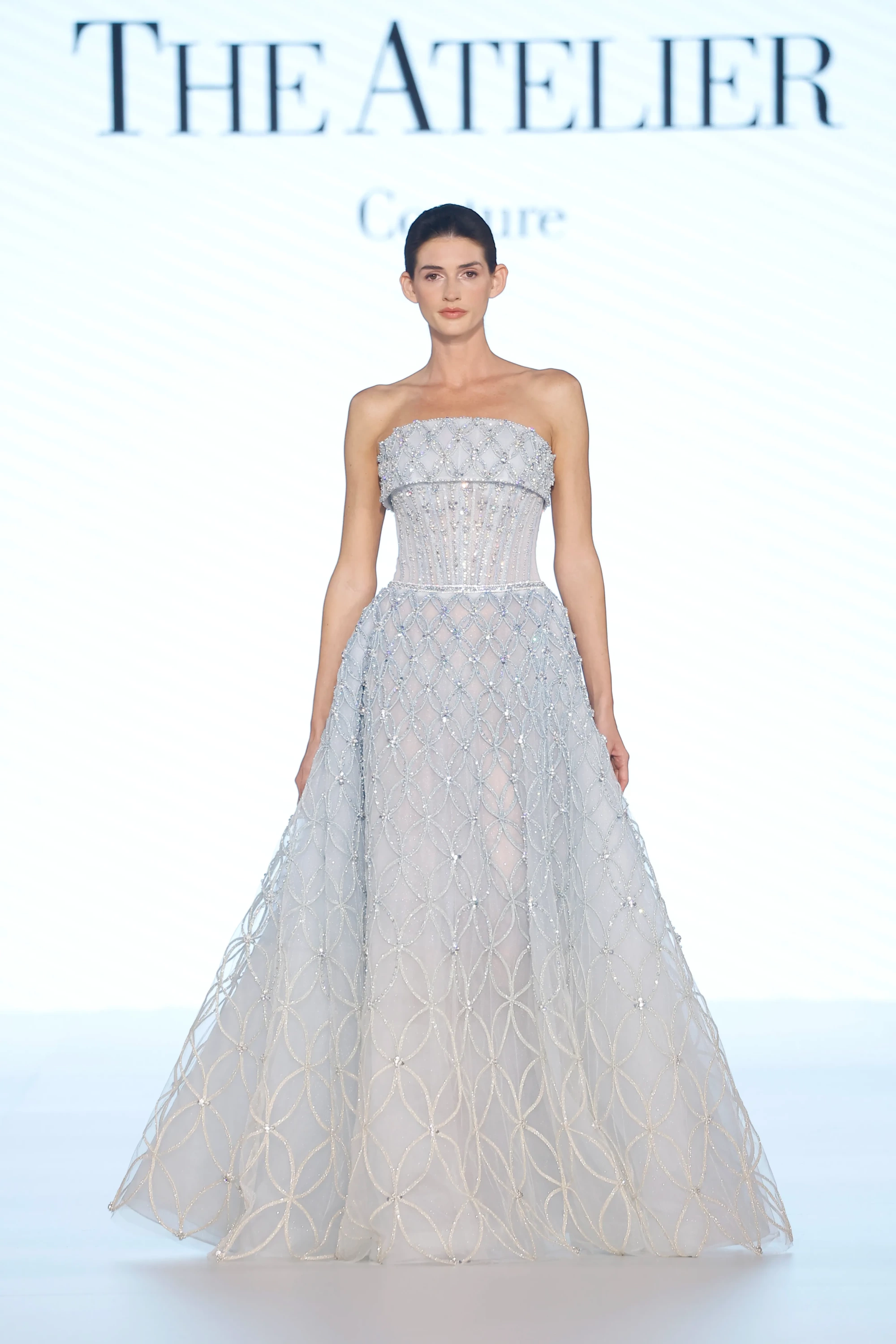 The Atelier İlkbahar 2027 Gelinlik Koleksiyonu'ndan, podyumda duran bir model, griye çalan mavi tonlarında, üst kısmı işlemeli ve Swarovski kristal detaylarla bezenmiş güçlü bir korsaja, etek kısmı ise ışıltılı geometrik desenlerle süslenmiş, katmanlı ve akışkan tül bir eteğe sahip olan modern bir gelinlik sunuyor.