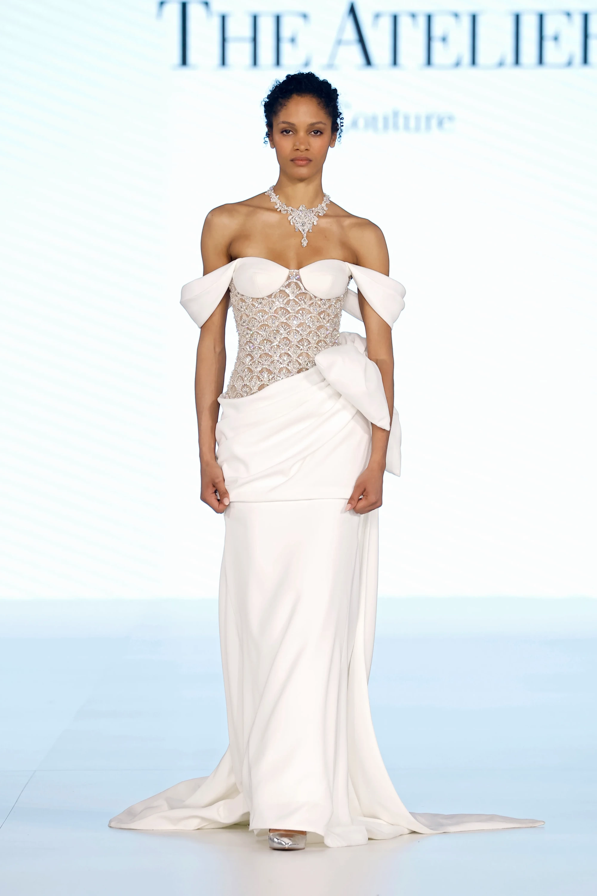 The Atelier by Prof. Jimmy Choo'nun 2027 gelinlik trendlerini yansıtan koleksiyonundan, podyumda duran esmer model, yoğun kristal işlemeli korsajı ve omuzları açıkta bırakan drapeli yaka detayıyla zarif, akışkan kumaşlı beyaz bir gelinlik içinde; belindeki şık drapeler ve gerdanındaki gösterişli taşlı kolyesiyle dikkat çekiyor.