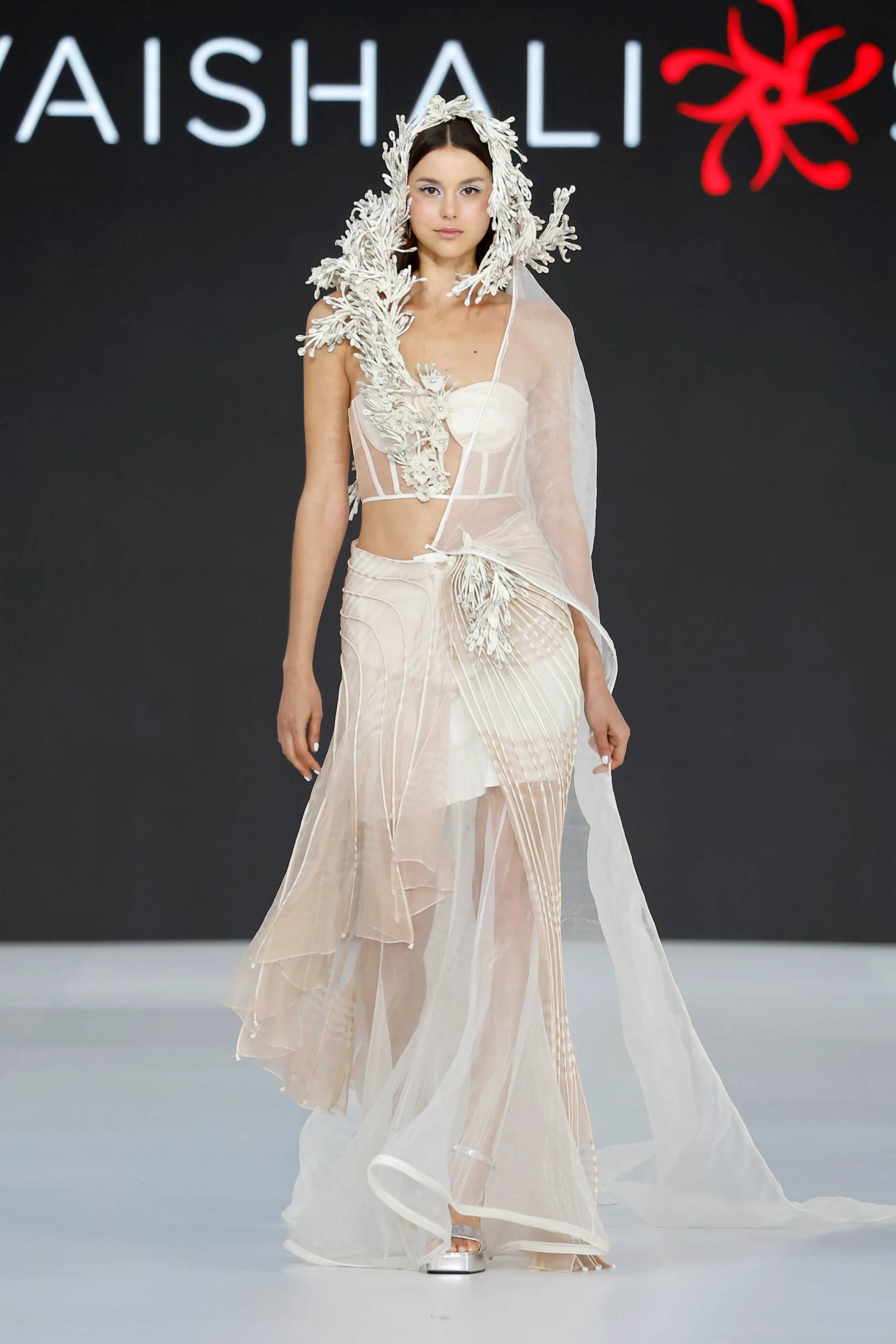 Model, Vaishali Studio 2027 İlkbahar Gelinlik Koleksiyonu'ndan, üst bedeni modern bir korseyle şekillendirilmiş, eteklerinde hareketli katmanlar, ince kordon detayları ve organik formlu üç boyutlu aplikeler barındıran zarif bir gelinlikle podyumda yürüyerek 2027 gelinlik trendlerinin özgün yorumunu sergiliyor.