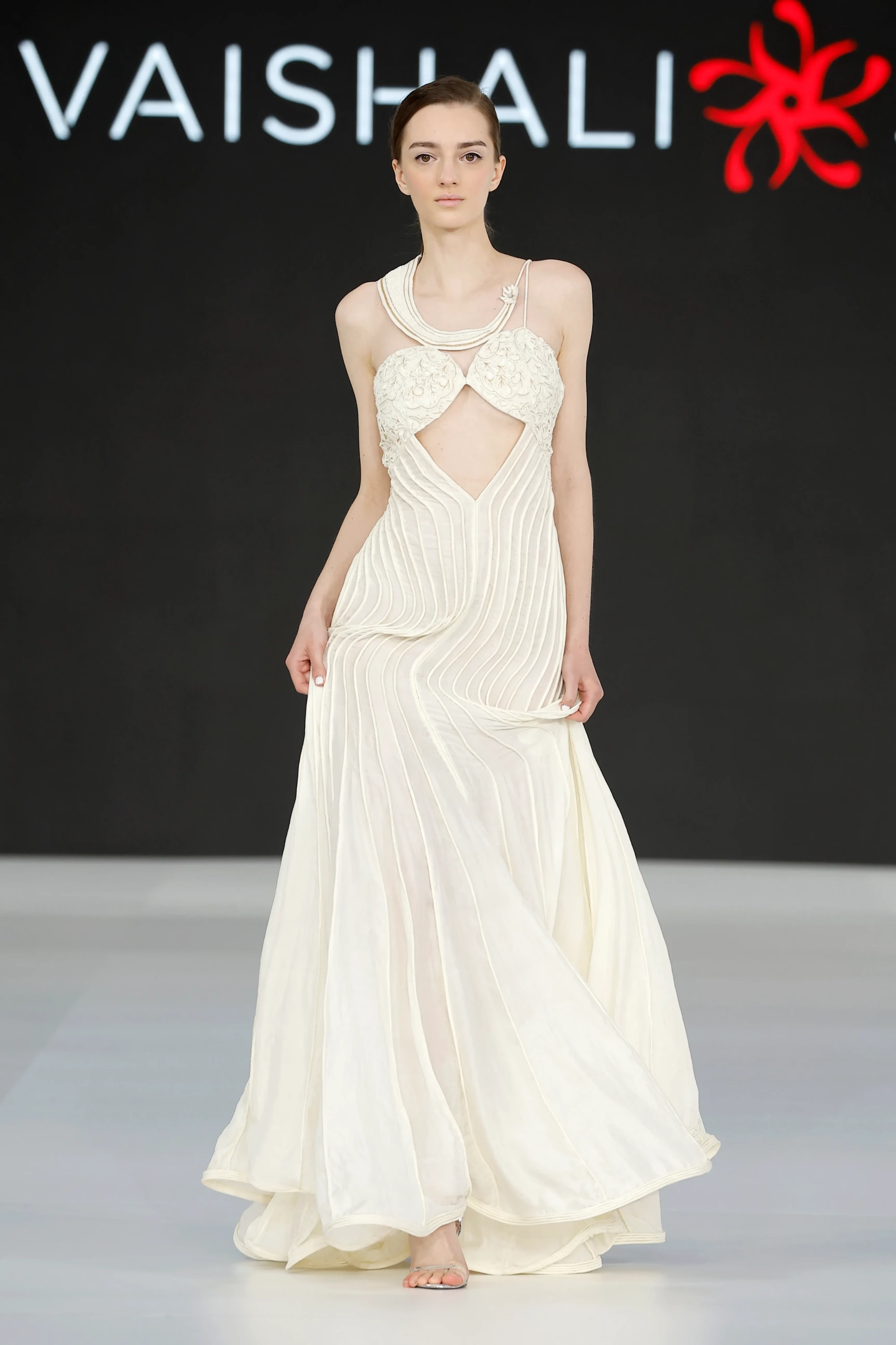 Barcelona Bridal Fashion Week 2026'da sergilenen Vaishali Studio'nun 2027 gelinlik trendlerini yansıtan, el işçiliği ip detaylarıyla süslenmiş boyun ve büst dekolteleri, karın bölgesindeki cesur kesimiyle dikkat çeken, dikey şeritlerle hareket ve akış kazanan fildişi rengi uzun gelinlikle podyumda yürüyen model.