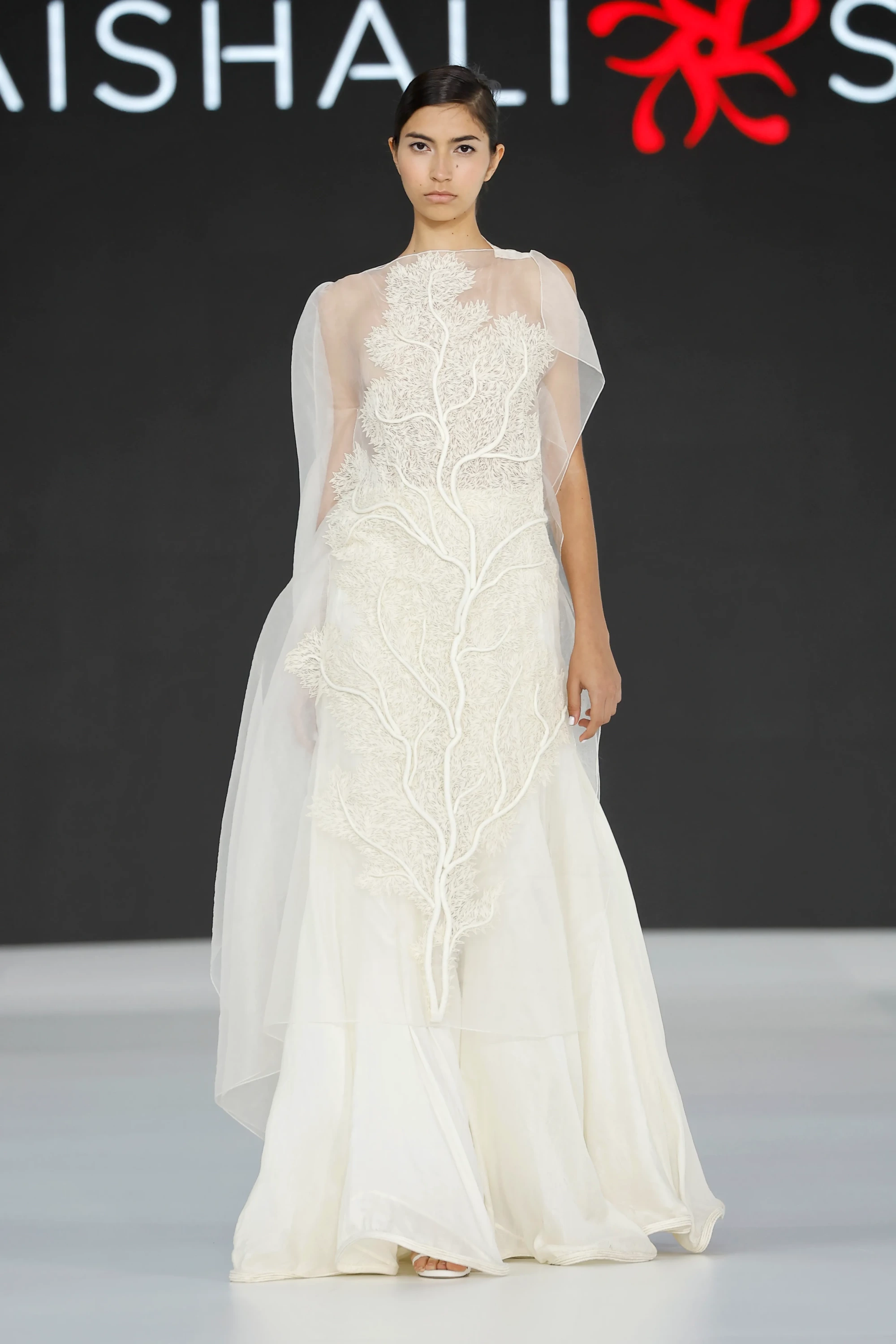 Model, siyah zeminli bir podyumda durmuş, Vaishali Studio'nun 2027 gelinlik trendlerini yansıtan, şeffaf katmanlı kumaşı ve gövdesinden eteğe uzanan, üç boyutlu dallı mercan formundaki krem rengi aplikeleriyle dikkat çeken, zarif ve akışkan bir gelinlik sergiliyor.