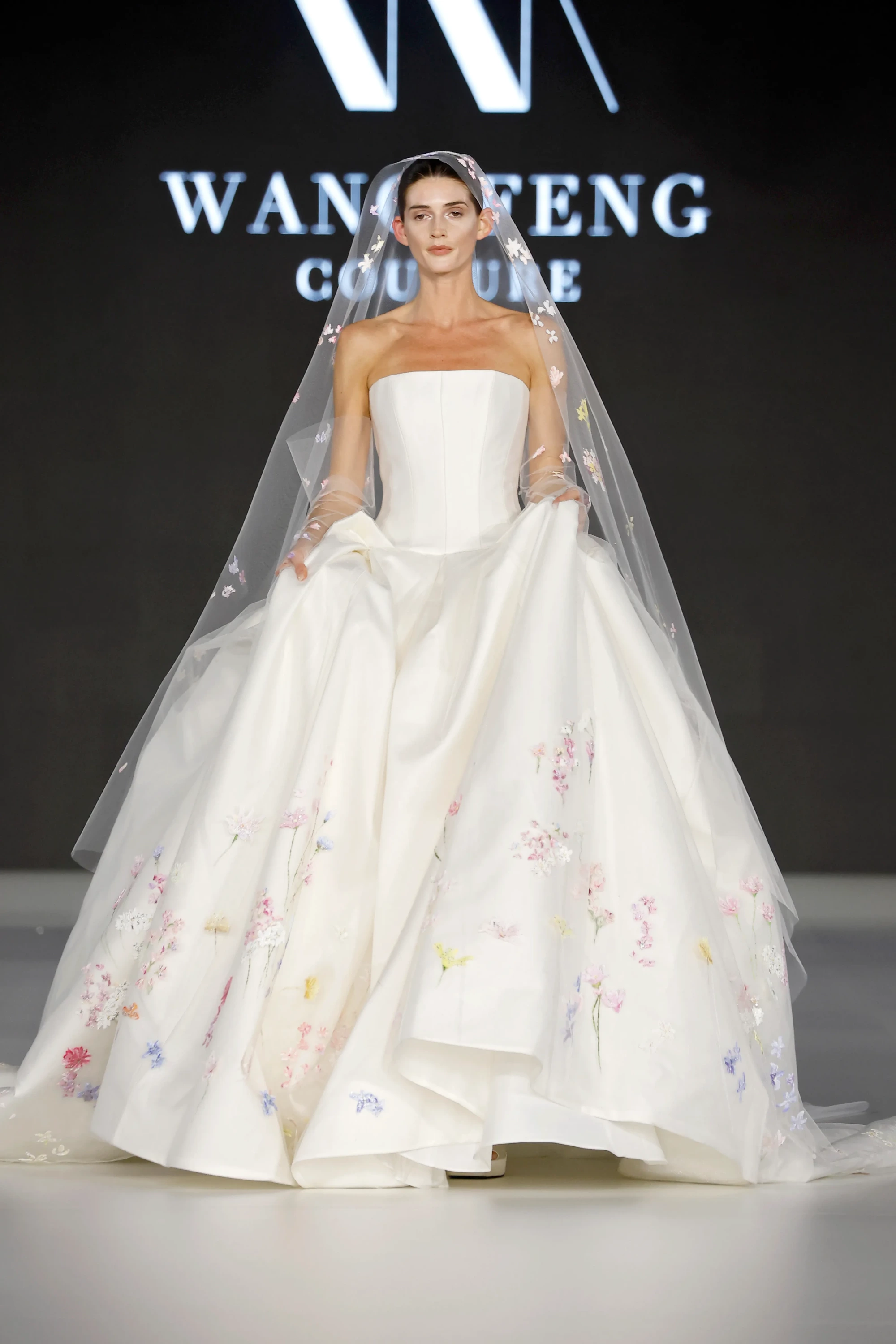 Wang Feng İlkbahar 2027 koleksiyonundan, modern gelinlik trendlerini yansıtan, uçuk pastel tonlardaki narin çiçek nakışlarıyla bezeli hacimli eteği ve straplez korsajıyla öne çıkan masalsı beyaz bir gelinlik giyen model, markanın logosunun önünde podyumda romantik bir duruş sergiliyor.