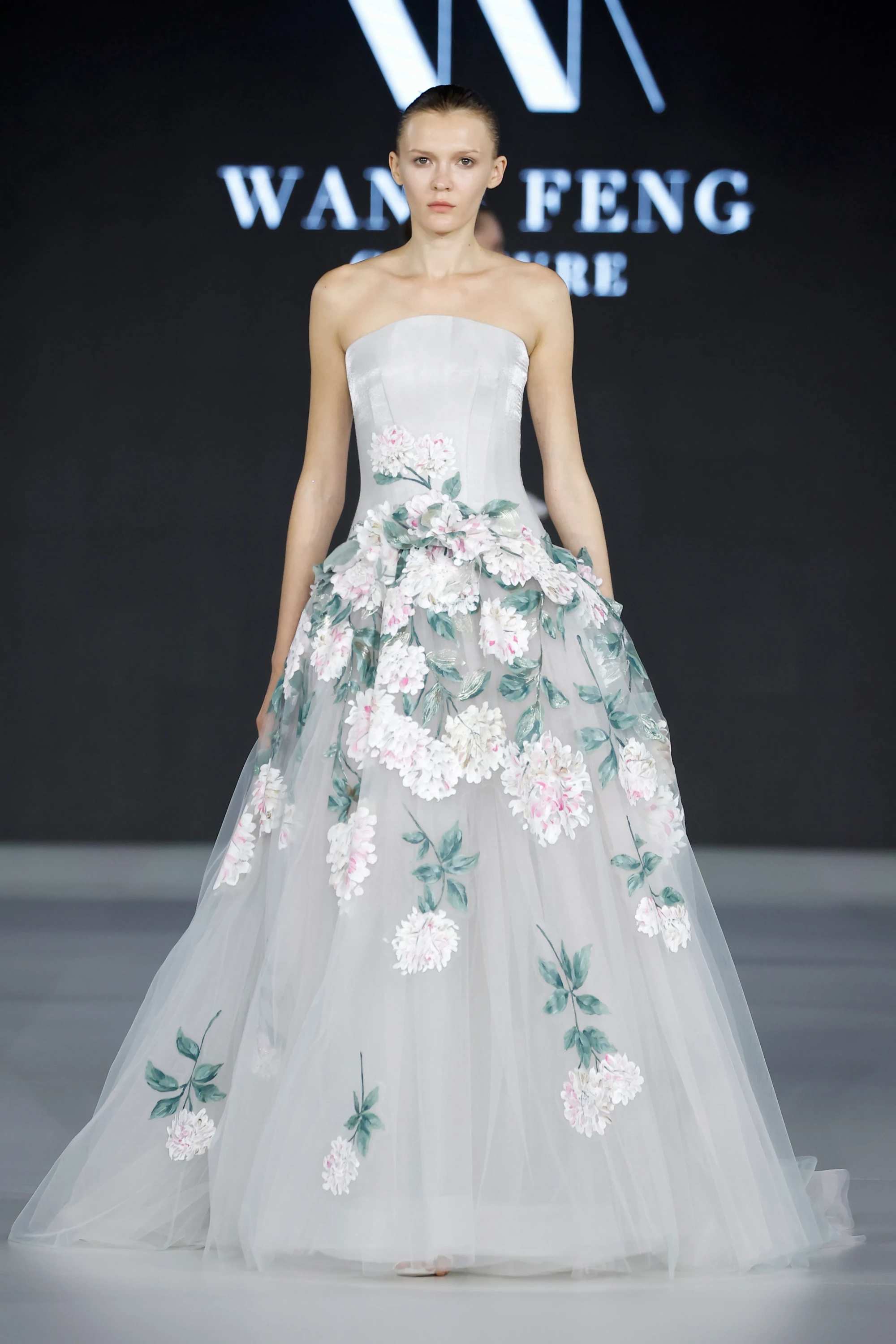 Podyumda tam boy görülen genç bir model, Wang Feng Couture'un İlkbahar 2027 koleksiyonundan, üst bedeni straplez ve oturaklı, etek kısmı ise kat kat tüllerle hacim kazanmış bembeyaz bir gelinlik içinde duruyor. Elbisenin etekleri, 2027 gelinlik trendleri arasında öne çıkan romantik 3D çiçek aplikeleri ve narin yeşil yaprak detaylarıyla süslenerek masalsı bir görünüm sunuyor.