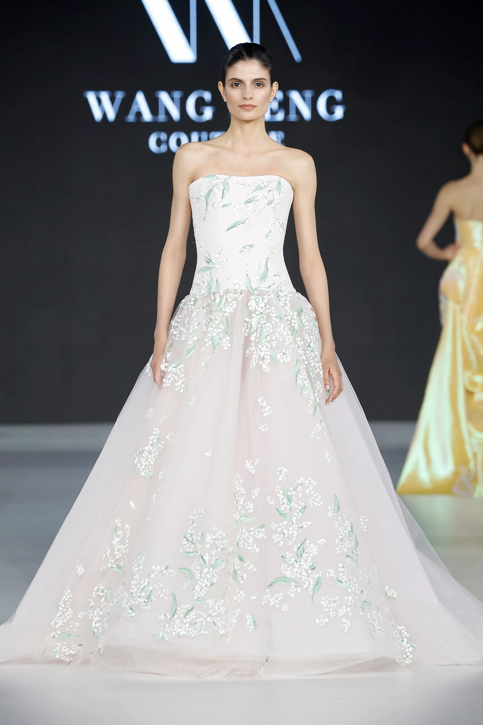 Barcelona Bridal Fashion Week 2026 podyumunda, Wang Feng'in İlkbahar 2027 gelinlik koleksiyonundan, zarif beyaz ve yeşil çiçek işlemeleriyle süslenmiş, straplez korsajlı üst ve kat kat tüllerden oluşan hacimli eteğiyle dikkat çeken bir gelinlik, 2027 gelinlik trendleri arasında yer alan masalsı ve romantik siluetleri yansıtıyor.