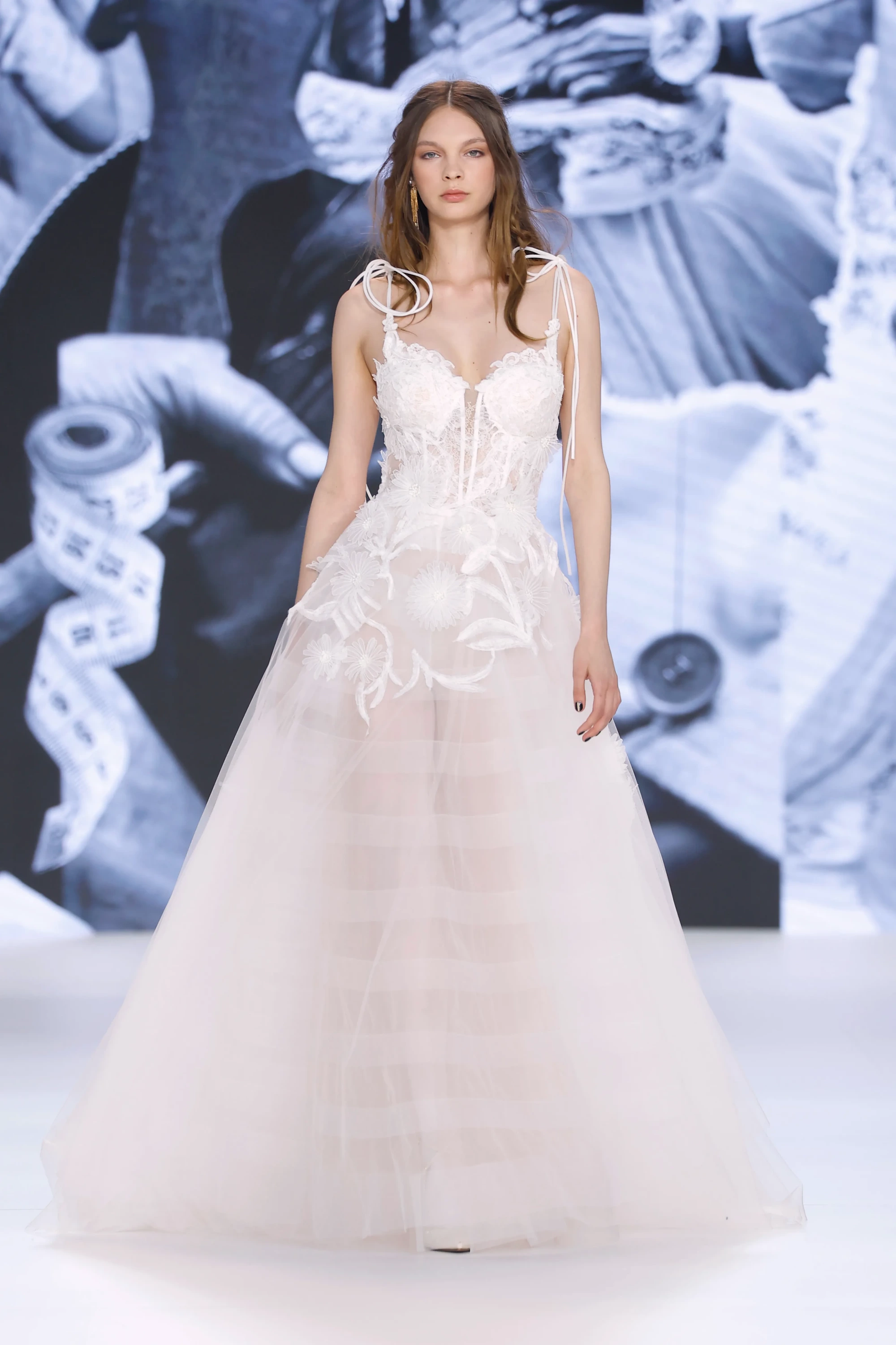 Barcelona Bridal Fashion Week'te sergilenen Yolancris 2027 İlkbahar Gelinlik Koleksiyonu'ndan bir parça olan bu tasarımda, transparan korsajlı üst beden omuzları bağcıklı detaylarla ve yoğun el işçiliği 3D çiçek aplikeleriyle zenginleşirken, hafif katmanlı tül etekler bohem ve modern romantizmi birleştiriyor.