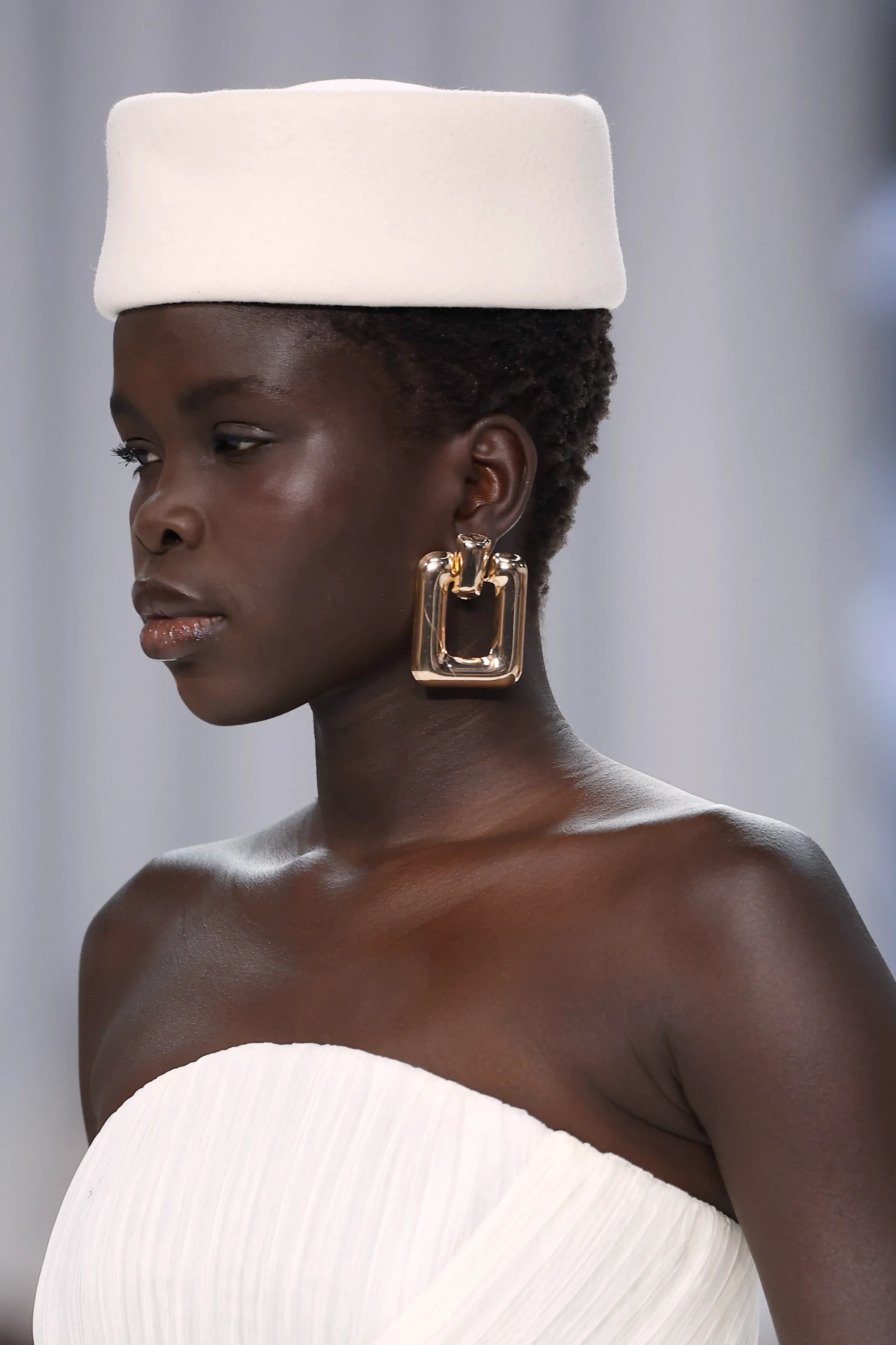 Barcelona Bridal Fashion Week 2026'dan Yolancris'in 2027 gelinlik koleksiyonunu tanıtan defilesinde, modern romantizm ve zanaatkârlığı yansıtan plili beyaz straplez bir elbiseyle, şık, kutu formunda bej bir şapka ve altın rengi kare küpeler takmış podyumda yürüyen modelin yakın çekim portresi.
