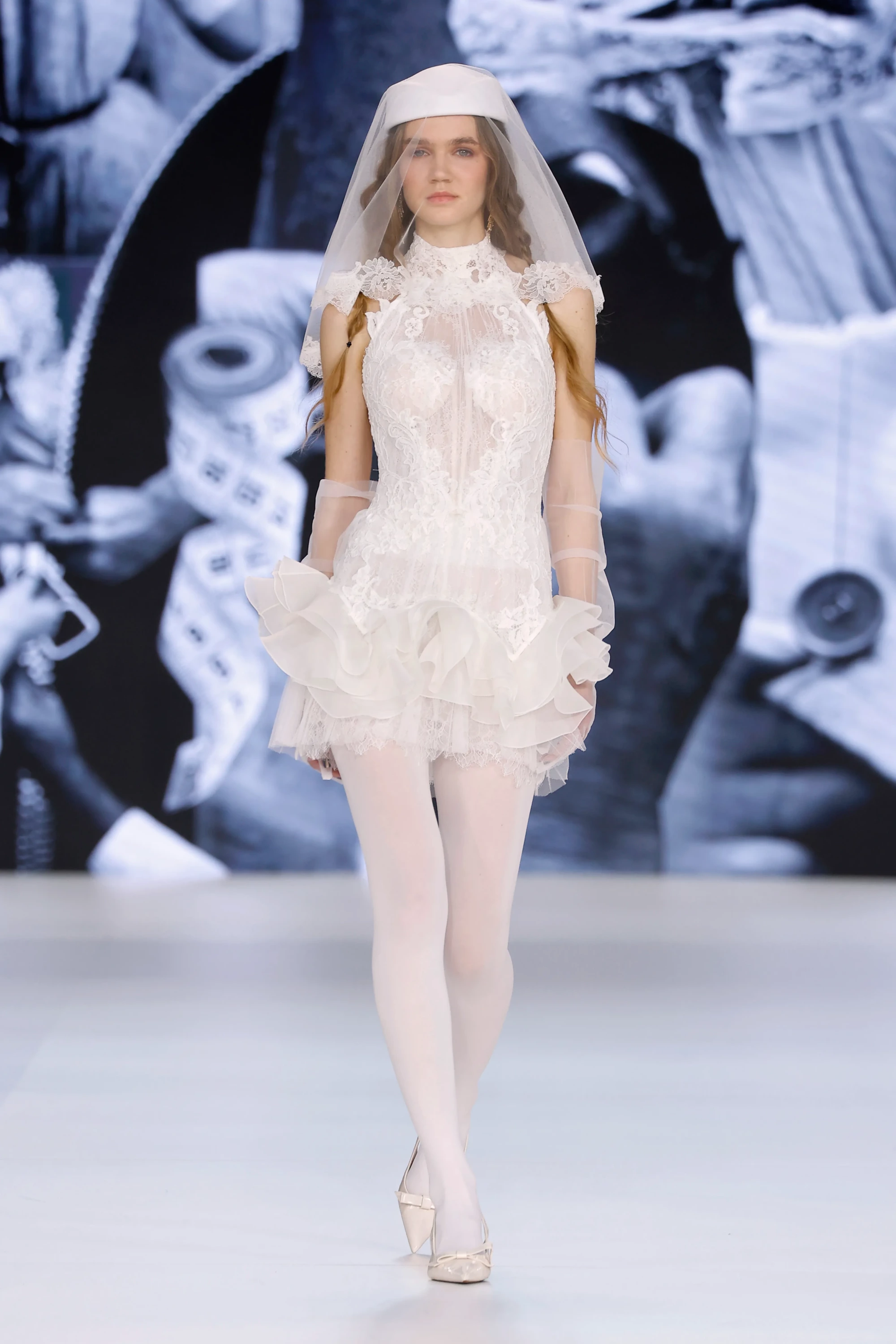 Barcelona Bridal Fashion Week 2026'dan Yolancris imzalı, yüksek yakalı, bol dantel detaylı ve transparan korsajlı kısa beyaz gelinlik, hacimli etek katmanları, tül eldivenleri ve şapka şeklindeki modern duvağıyla 2027 gelinlik trendlerinin özgün ve cesur bir yorumunu sunuyor.