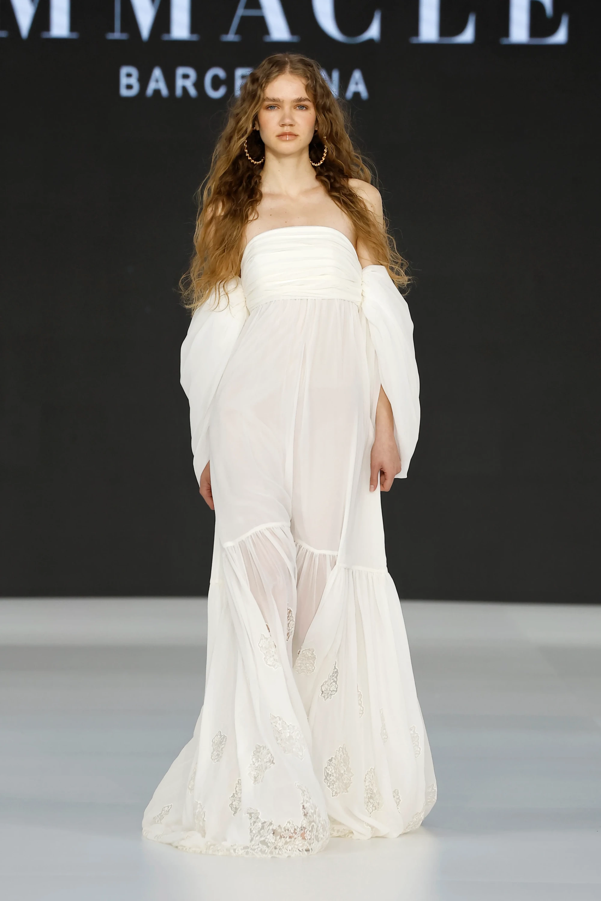 Barcelona Bridal Fashion Week 2026’da Immaclé defilesinde podyumda yürüyen model, straplez formda, transparan şifon katmanlı ve dantel işlemeli beyaz gelinlik giyiyor; düşük omuzlu balon kollu detaylara sahip tasarım, uçuşan katmanlı etekle tamamlanırken model uzun dalgalı saçları ve halka küpeleriyle doğal bir görünüm sergiliyor, arka planda Immaclé logosu yer alıyor.