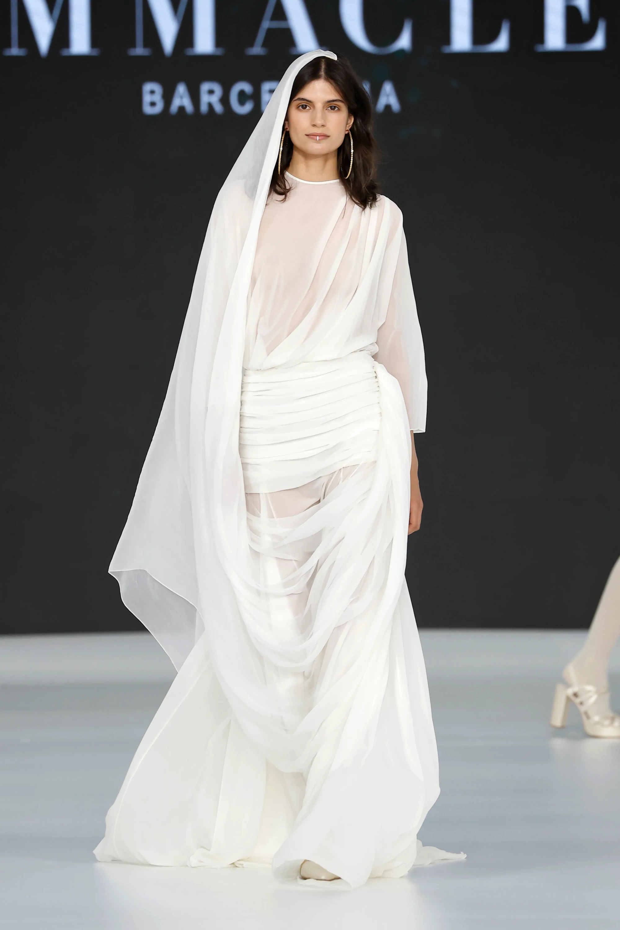 Barcelona Bridal Fashion Week 2026’da Immaclé defilesinde podyumda yürüyen model, transparan katmanlı ve drapeli detaylara sahip uzun beyaz gelinlik giyiyor; ince tül kumaşın vücut üzerinde katlanarak şekillendiği tasarım, uzun duvak, asimetrik etek formu ve sade aksesuarlarla tamamlanırken arka planda Immaclé logosu yer alıyor.