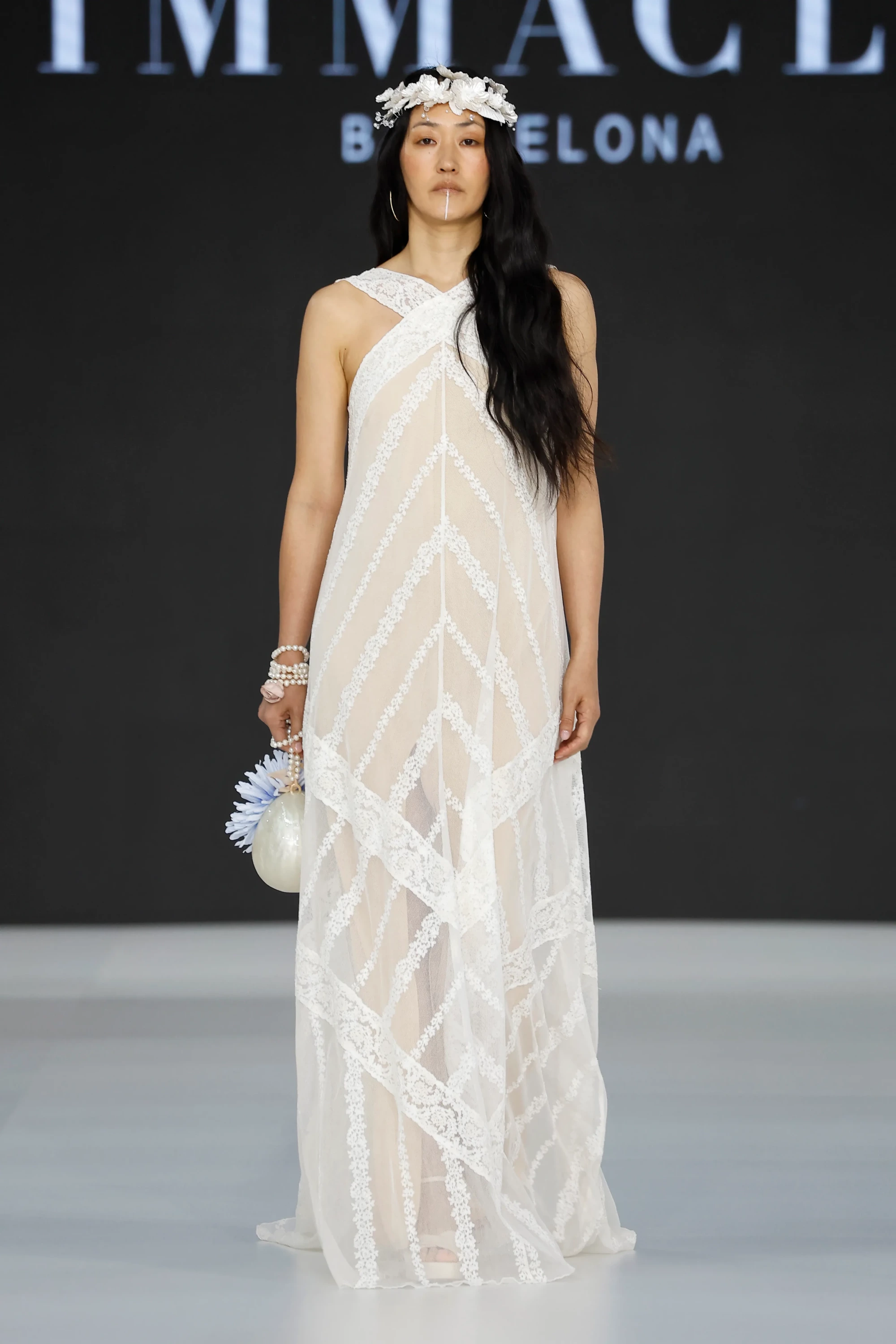 Barcelona Bridal Fashion Week 2026’da Immaclé defilesinde podyumda yürüyen model, çapraz askılı, transparan tül üzerine geometrik dantel işlemeli uzun beyaz gelinlik giyiyor; düz kesimli akışkan silüete sahip tasarım, saçta çiçekli taç, bilekte inci bilezikler ve elde çiçek detaylı küçük çanta ile tamamlanırken arka planda Immaclé logosu yer alıyor.
