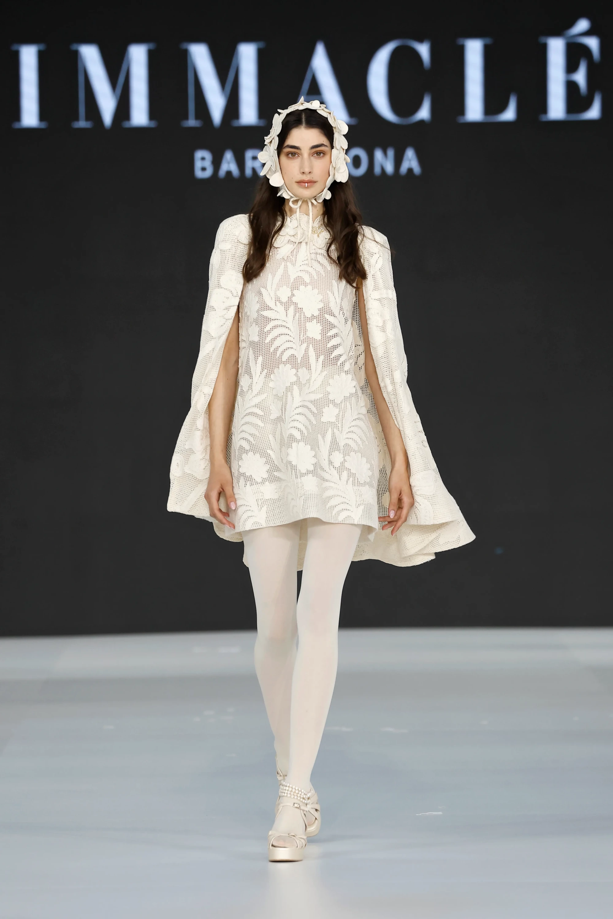 Barcelona Bridal Fashion Week 2026’da Immaclé defilesinde podyumda yürüyen model, yüksek yakalı, uzun kollu ve pelerin etkili kesime sahip kısa beyaz gelinlik giyiyor; dantel işlemeli mini elbise transparan çorap ve bantlı ayakkabılarla tamamlanırken model çiçek detaylı başlık aksesuarı kullanıyor, arka planda Immaclé logosu yer alıyor.