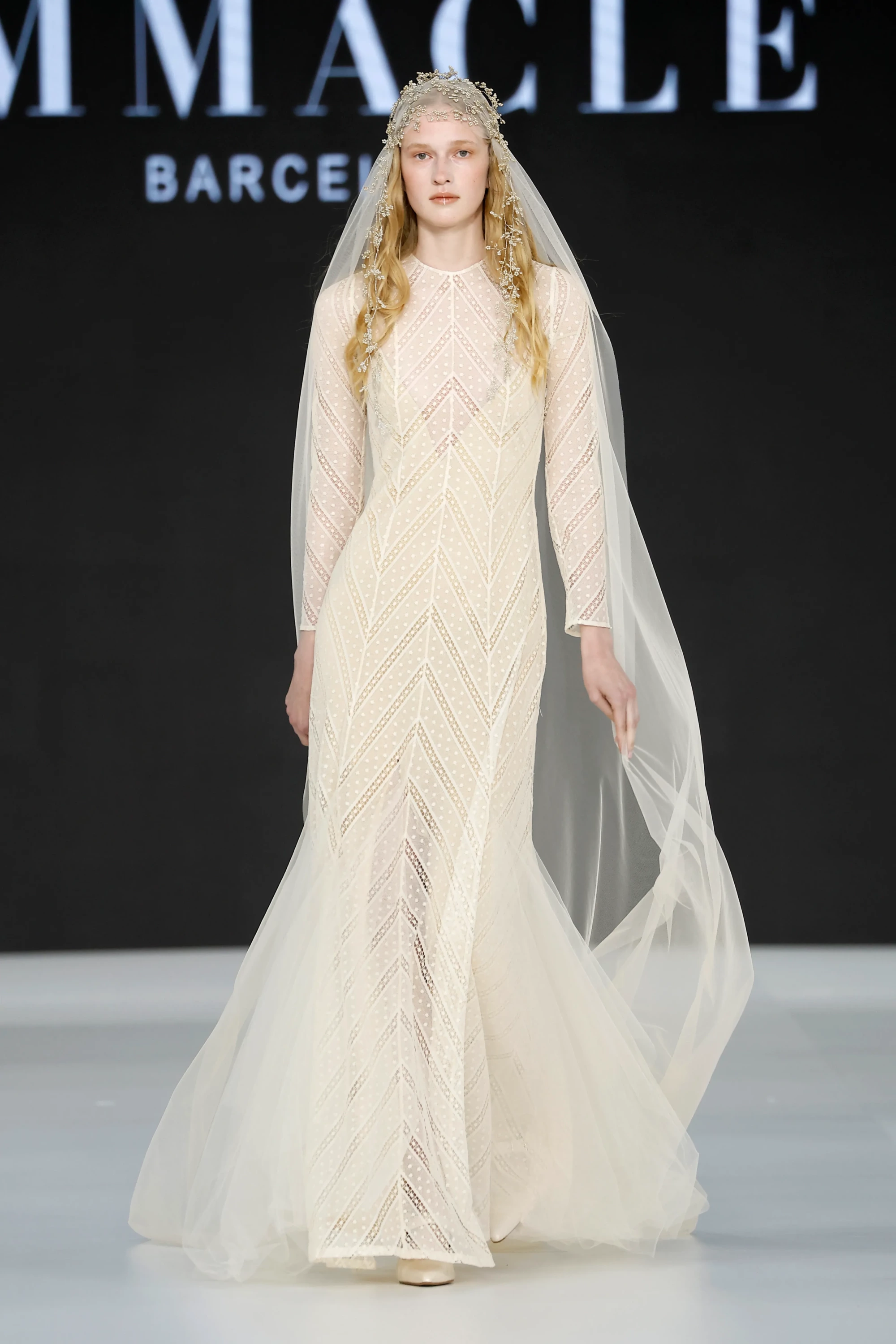 Barcelona Bridal Fashion Week 2026’da Immaclé defilesinde podyumda yürüyen model, uzun kollu, yüksek yakalı ve vücuda oturan kesime sahip transparan dantel işlemeli krem tonlarında gelinlik giyiyor; geometrik desenli kumaş detaylarıyla öne çıkan tasarım, uzun tül duvak ve çiçek işlemeli başlıkla tamamlanırken arka planda Immaclé logosu yer alıyor.