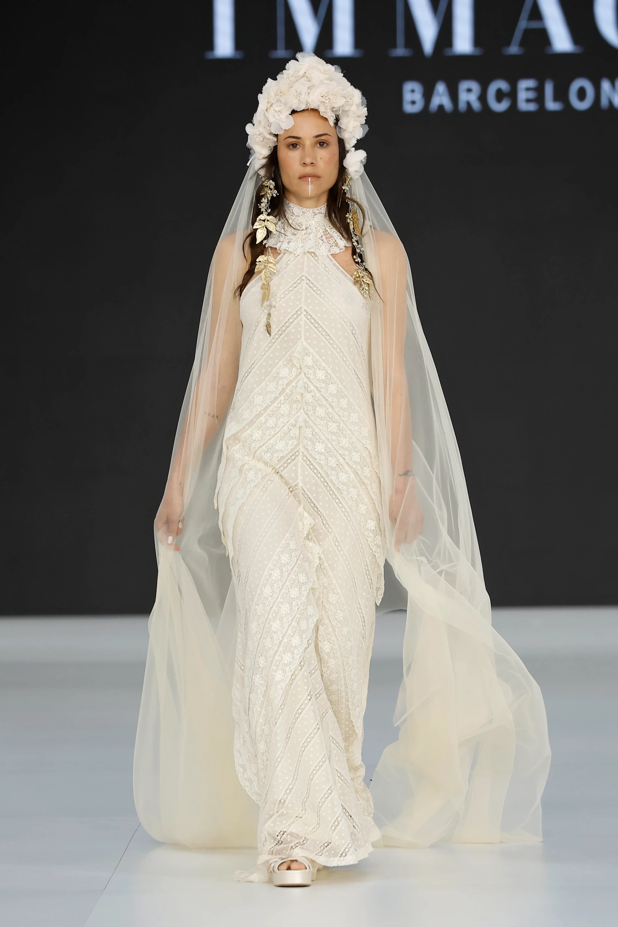 Barcelona Bridal Fashion Week 2026’da Immaclé defilesinde podyumda yürüyen model, yüksek yakalı, transparan detaylı ve yoğun dantel işlemeli uzun beyaz gelinlik giyiyor; katmanlı dantel dokulara sahip tasarım, uzun tül duvak, çiçekli hacimli başlık ve altın tonlu aksesuarlarla tamamlanırken arka planda Immaclé logosu yer alıyor.