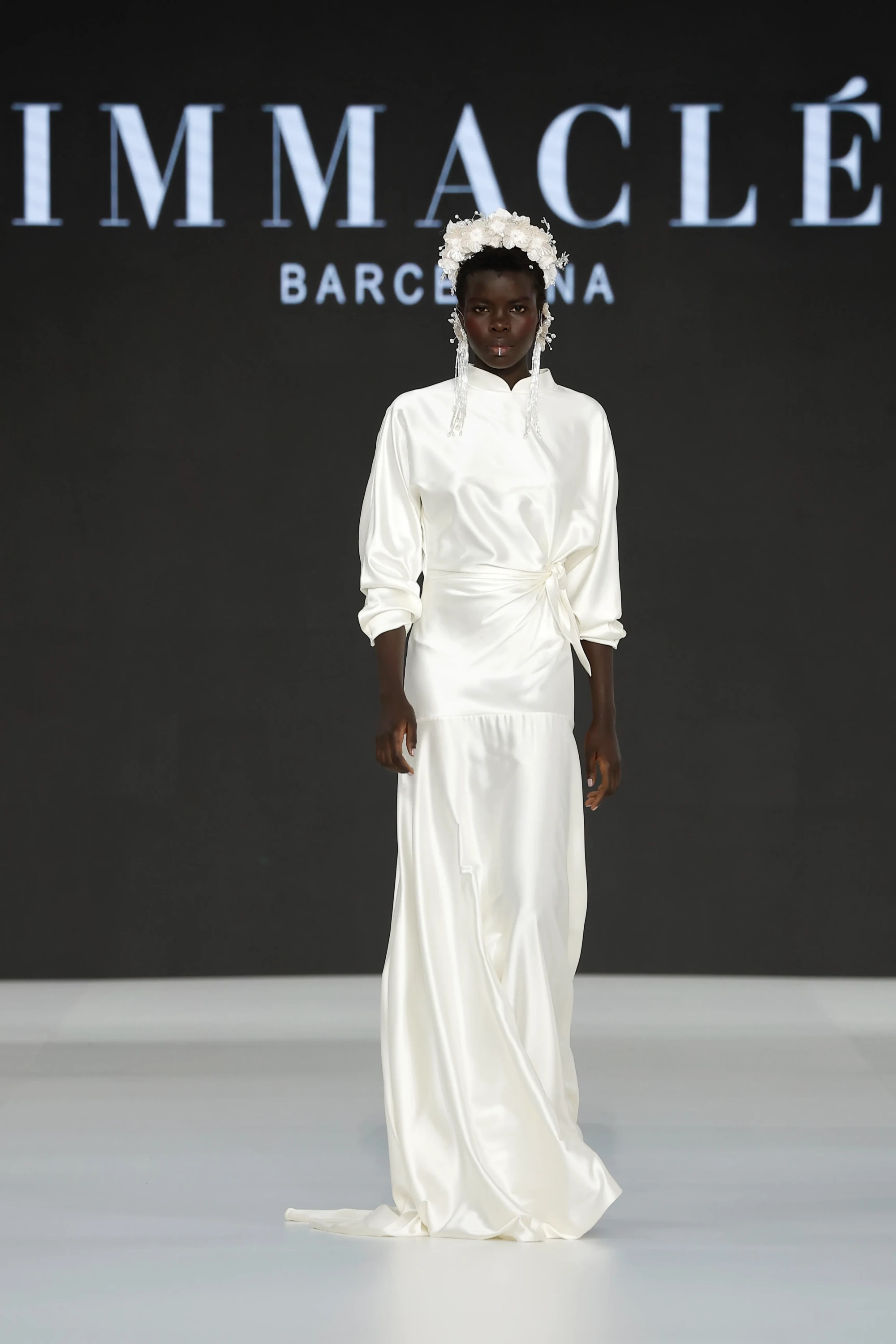 Barcelona Bridal Fashion Week 2026’da Immaclé defilesinde podyumda yürüyen model, uzun kollu, yüksek yakalı, ipeksi saten dokulu beyaz gelinlik giyiyor; bel kısmında drapeli bağlama detayı bulunan tasarım, düz kesim uzun etekle tamamlanırken model saçında çiçekli aksesuar ve sarkan inci detaylı başlık taşıyor, arka planda Immaclé logosu yer alıyor.
