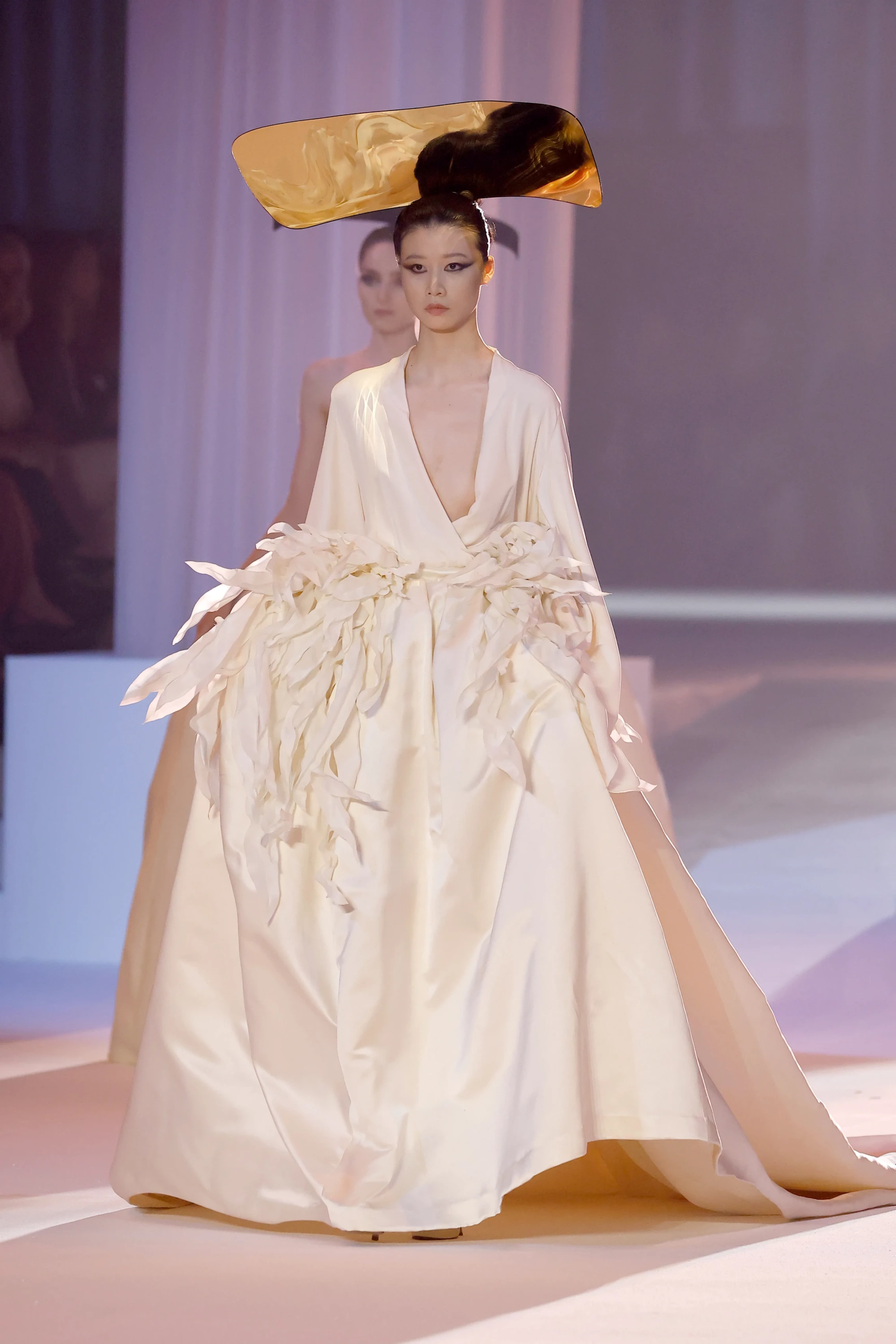 Stéphane Rolland'ın Barcelona Bridal Night'taki 2027 İlkbahar gelinlik koleksiyonundan, başındaki büyük, altın rengi heykelsi aksesuar ve belindeki dramatik kumaş işlemeleriyle dikkat çeken, derin V yakalı, akışkan krem rengi bir gelinlikle podyumda yürüyen bir model, haute couture gelin modasına mimari bir yorum getiriyor.