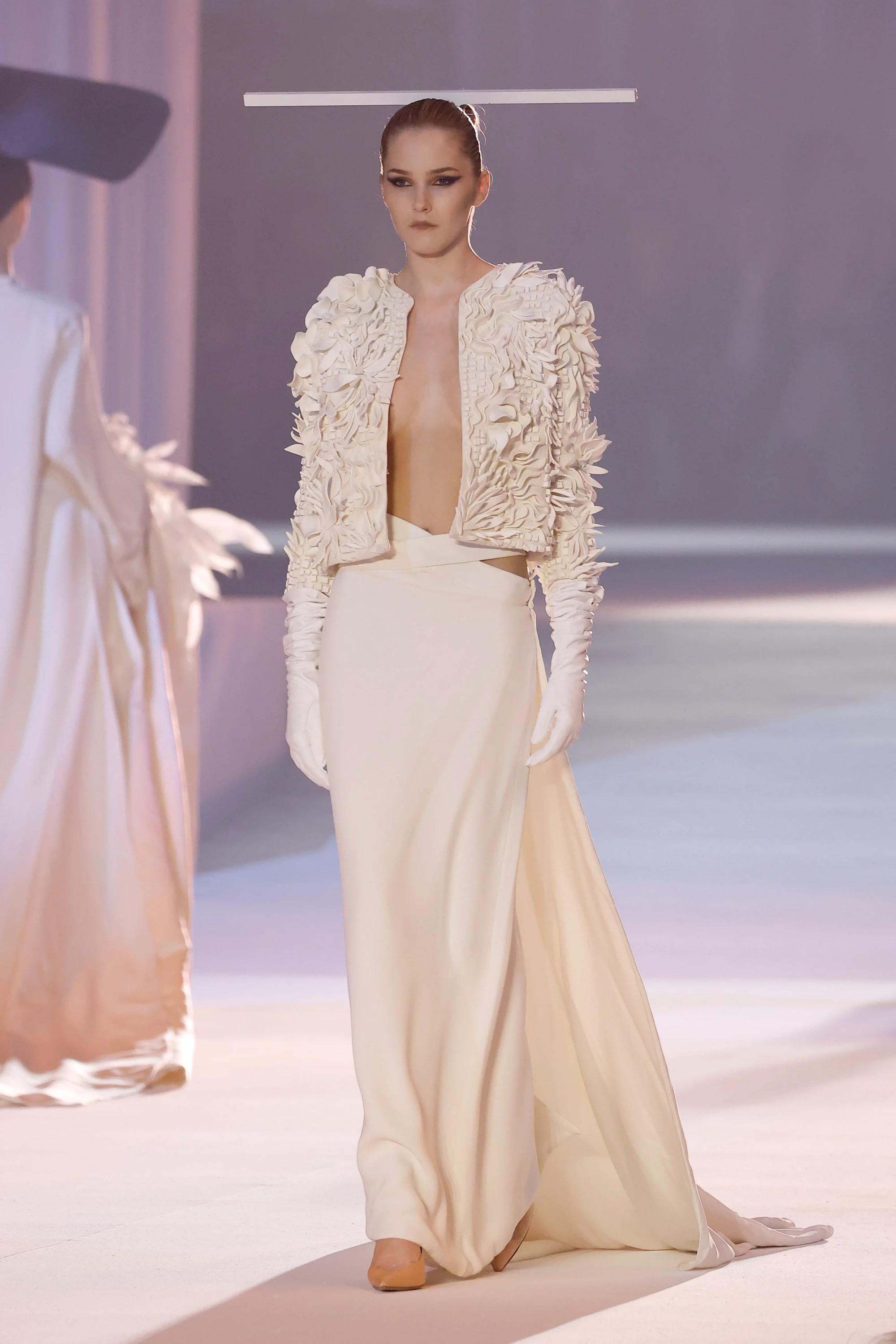Stéphane Rolland'ın Barcelona Bridal Night'ta sergilediği 2027 gelinlik modası tasarımlarından, pistte yürüyen, başındaki yatay mimari aksesuarı, 3D çiçek aplikeli kısa ceketi, uzun eldivenleri ve derin yırtmaçlı akışkan krem rengi eteğiyle heykelsi bir zarafet sunan model.