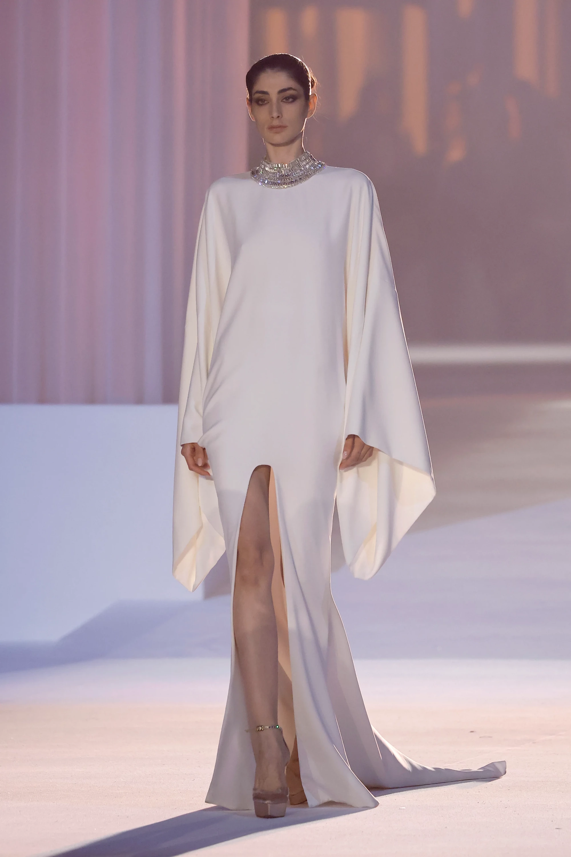Stéphane Rolland'ın Barcelona Bridal Night'ta sergilediği 2027 İlkbahar gelinlik koleksiyonundan, geniş ve akışkan kolları, yüksek bacak yırtmacı ve göz alıcı kristal yakalı ekru bir gelinliği taşıyan model, podyumda heykelsi bir zarafetle yürüyor.