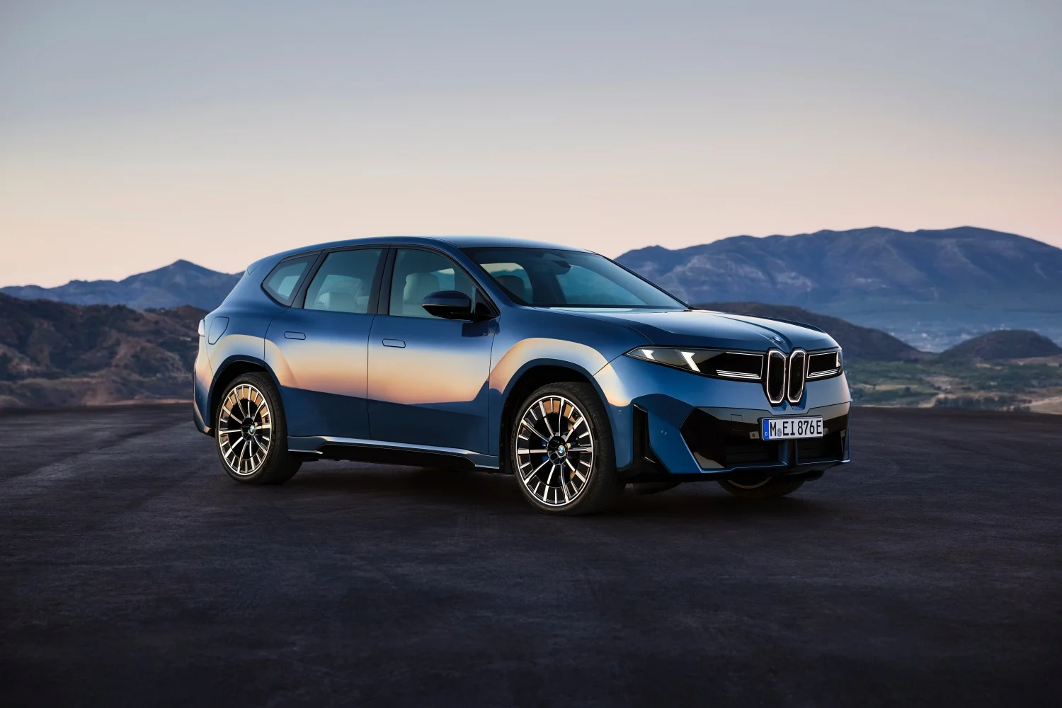 Alacakaranlıkta dağlık bir manzara önünde duran koyu mavi renkli BMW elektrikli lüks SUV, yan tarafında gün batımı renklerini yansıtırken, Neue Klasse tasarım dilini ve modern far detaylarını net bir şekilde gösteriyor.