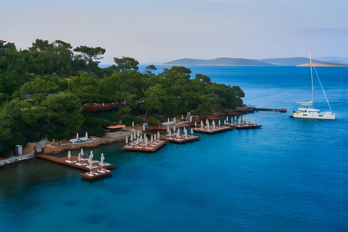 Bodrum Loft'un turkuaz Ege Denizi'ne uzanan ahşap iskeleleri ve yeşilliklerle çevrili kıyısı, beyaz şemsiyeli şezlongları ve sakin koyda demirlemiş zarif katamaranıyla, sade ve lüks bir Bodrum düğün deneyimi için büyüleyici bir manzara sunuyor.