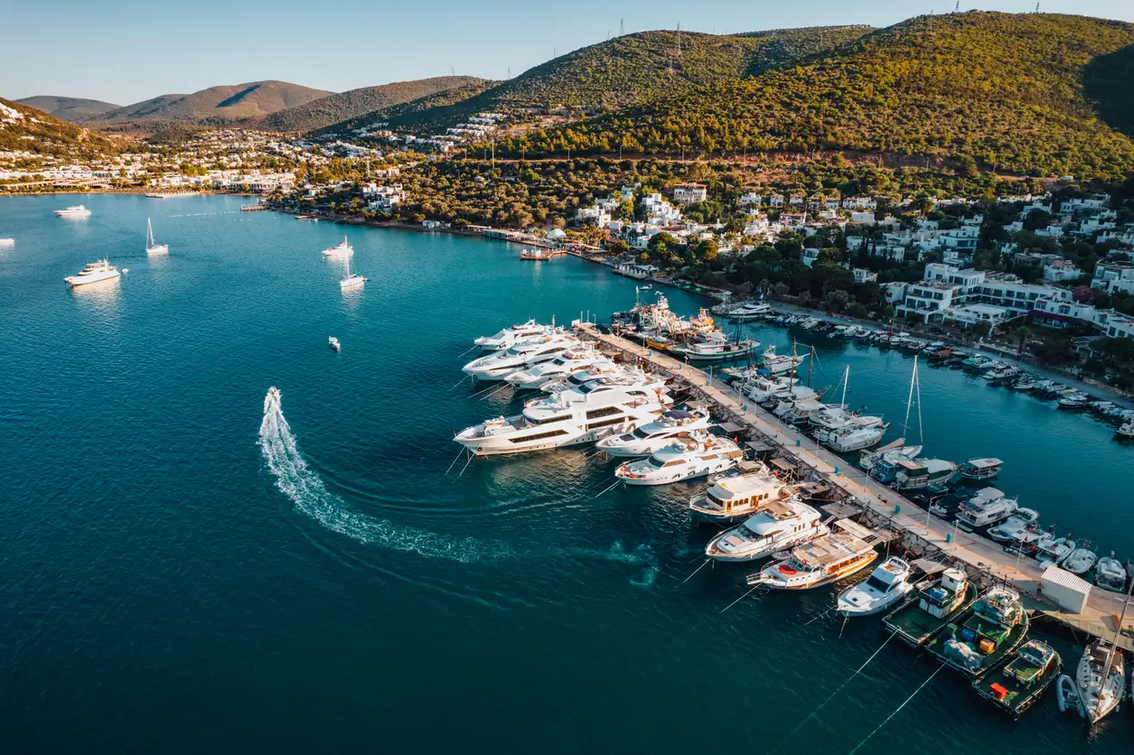 Bodrum'un Torba Koyu'nun havadan görünümünde, berrak turkuaz sularda demirli çok sayıda beyaz yat ve tekneyle dolu uzun bir marina iskele hattı, yemyeşil tepelerle çevrili kıyı yapıları ve denizde iz bırakan bir sürat teknesi görülüyor.