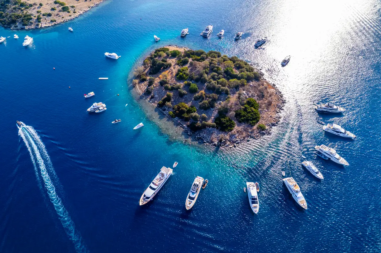 Bodrum Türkbükü'nün kristal berraklığındaki turkuaz sularında, yemyeşil bitki örtüsüyle kaplı küçük bir adanın etrafına demirlemiş çok sayıda lüks yatın ve teknenin havadan çekilmiş nefes kesen bir görüntüsü, bir teknenin arkasında bıraktığı köpüklü izle dikkat çekiyor.
