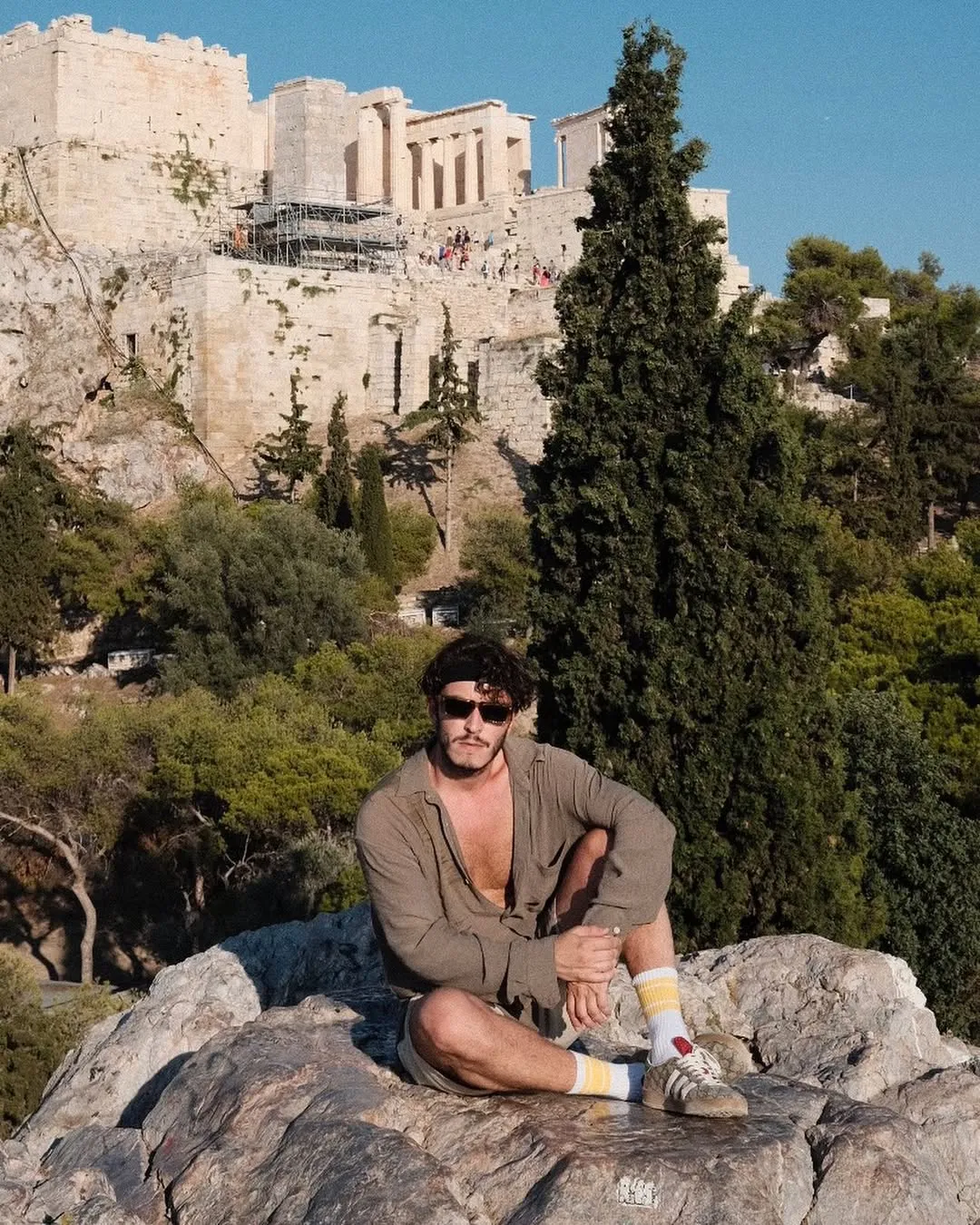Boran Kuzum, kahverengi güneş gözlükleri ve günlük kıyafetleriyle, arkasında antik Atina Akropolisi'nin görkemli Parthenon'u ve diğer yapıları ile ağaçlarla kaplı bir tepede kayalık bir zeminde rahatça oturuyor.