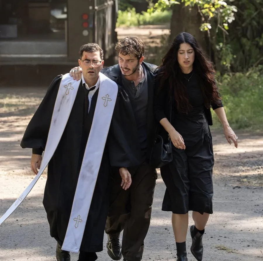 Boran Kuzum, Netflix’in Big Mistakes dizisinden bir sahnede, Yusuf karakteri olarak, siyah cübbeli ve beyaz stola takmış, bitkin görünen bir adamın omzuna kolunu atmış, yanında ciddi ifadeyle yürüyen uzun saçlı bir kadınla ağaçlık bir alanda toprak yolda endişeli ve koruyucu bir duruş sergiliyor.