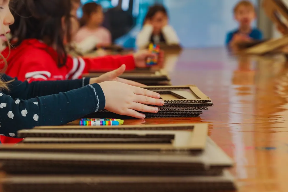 Ön planda, üzerinde beyaz desenler olan koyu mavi uzun kollu bir tişört giyen bir çocuğun elleri, parlak ahşap bir masa üzerindeki karton çerçevelerden oluşan bir yığının üzerinde duruyor; yanındaki rengarenk boya kalemleri ve arkadaki bulanık diğer çocuklar, 23 Nisan Çocuk Bayramı atölyesi etkinliğini yansıtıyor.