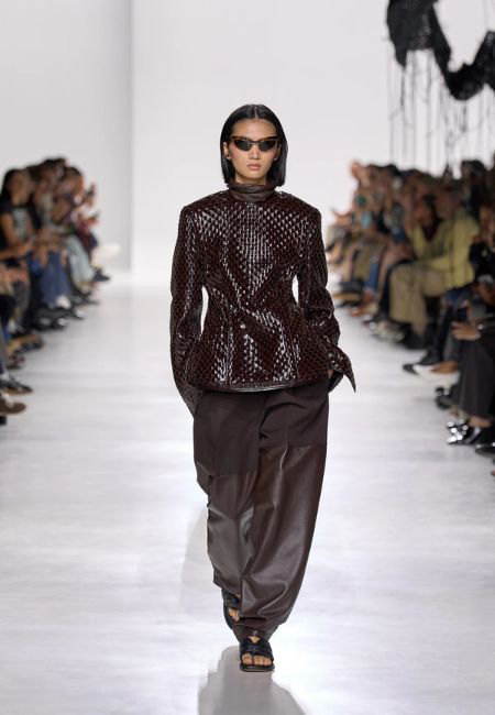 Bottega Veneta'nın 2026 İlkbahar-Yaz koleksiyonundan bir model, podyumda koyu bordo rengi, kabartmalı dokulu, omuzları yapılı bir ceket ve bol kahverengi pantolon giyerek yürüyor; bu kıyafet güçlü siluetler ve dikkat çekici dokusal yüzeyler trendini vurguluyor.