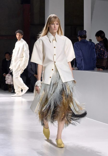 Bottega Veneta defilesinden bir model, 2026 ilkbahar-yaz moda trendleri arasında yer alan dikkat çekici dokuları ve dinamik yüzeyleri sergileyen, ekru kısa kollu gömlek, gri, altın, kahverengi ve siyah tonlarında uzun püsküllü etek ve açık sarı ayakkabılar giyerek parlak podyumda yürüyor.