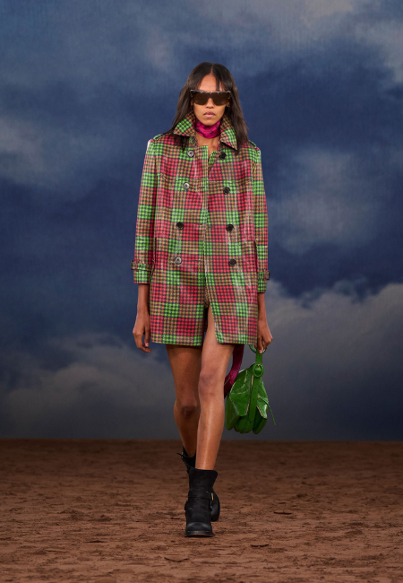Burberry'nin 2026 ilkbahar-yaz defilesinden, koyu renk güneş gözlüğü ve pembe fular takmış model, canlı kırmızı, yeşil ve kahverengi ekoseli, kısa kesimli, kruvaze trençkotuyla dönemin moda trendlerini yansıtan özgün bir siluet sunuyor; kombini yeşil el çantası ve siyah botlarla tamamlıyor, bulutlu bir gökyüzü altında toprak zeminde yürüyor.