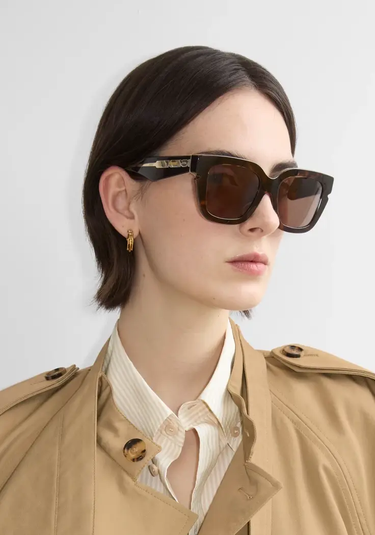 Bir kadın, koyu kahverengi kaplumbağa kabuğu desenli, geniş ve köşeli çerçeveli Burberry güneş gözlüğü takmış, saplarında dikkat çekici altın detaylar bulunan bu gözlüklerle bej bir trençkot ve açık renk çizgili gömleğiyle şık bir duruş sergiliyor, kısa kahverengi saçları ve altın halka küpesiyle zarif bir yan profilden poz veriyor.