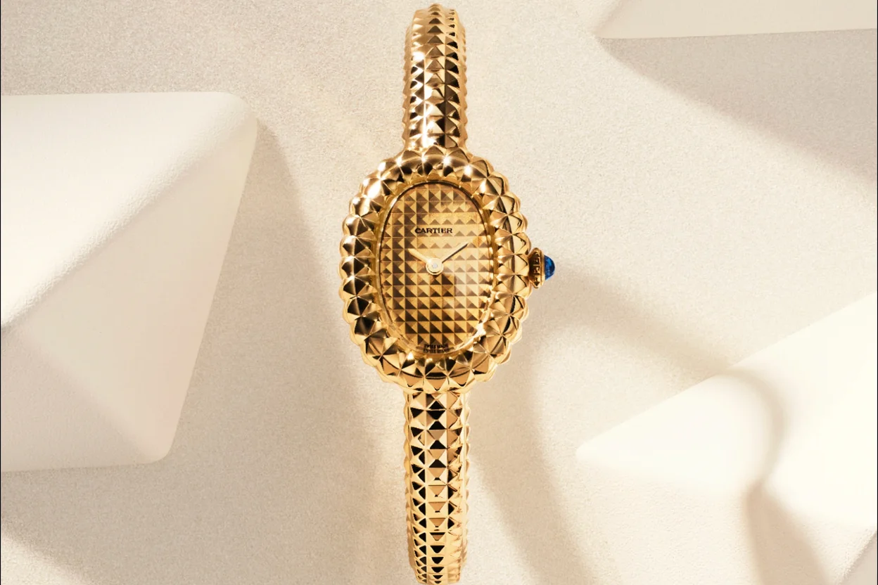 Açık renk minimalist bir zemin üzerinde dikey olarak konumlandırılmış, oval kasa ve kordonu üç boyutlu piramit benzeri geometrik desenlerle süslenmiş, sağ tarafında mavi cabochon safir kurma kolu bulunan, zarif Cartier Baignoire sarı altın kadın saati.